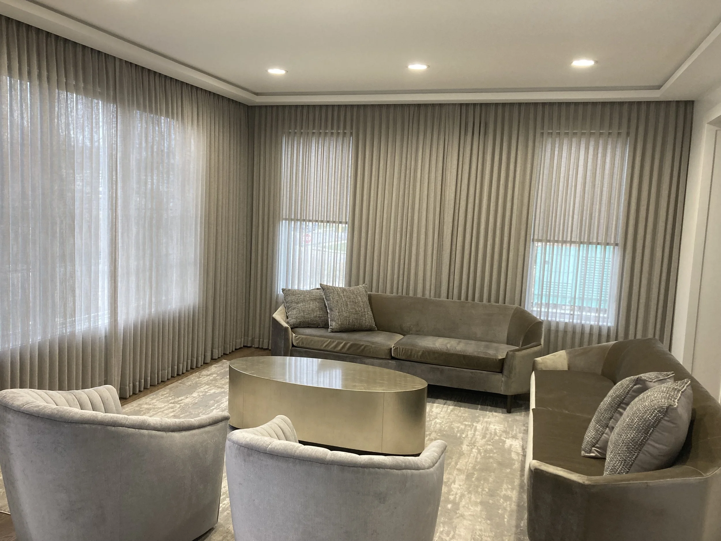Living room with beige and gray furniture, including a sofa, armchairs, a coffee table, and large windows with beige curtains.