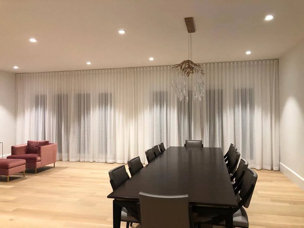 Empty dining room with black table and eight chairs, pink armchair and ottoman, white curtains, chandelier, and recessed ceiling lights.