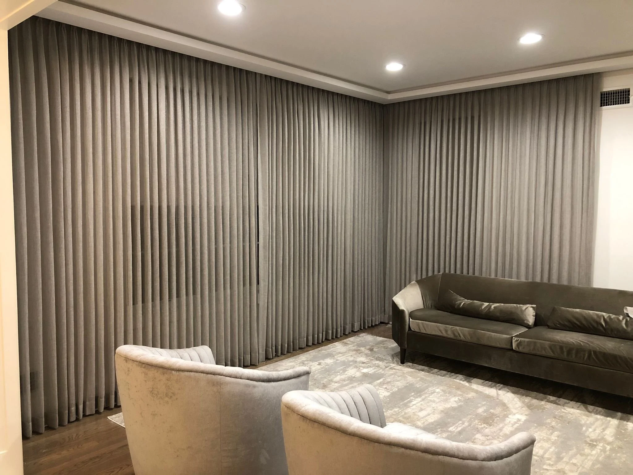 Living room with beige velvet armchairs, a dark green velvet sofa with throw pillows, gray vertical curtains covering the windows, ceiling lights, and a patterned area rug.
