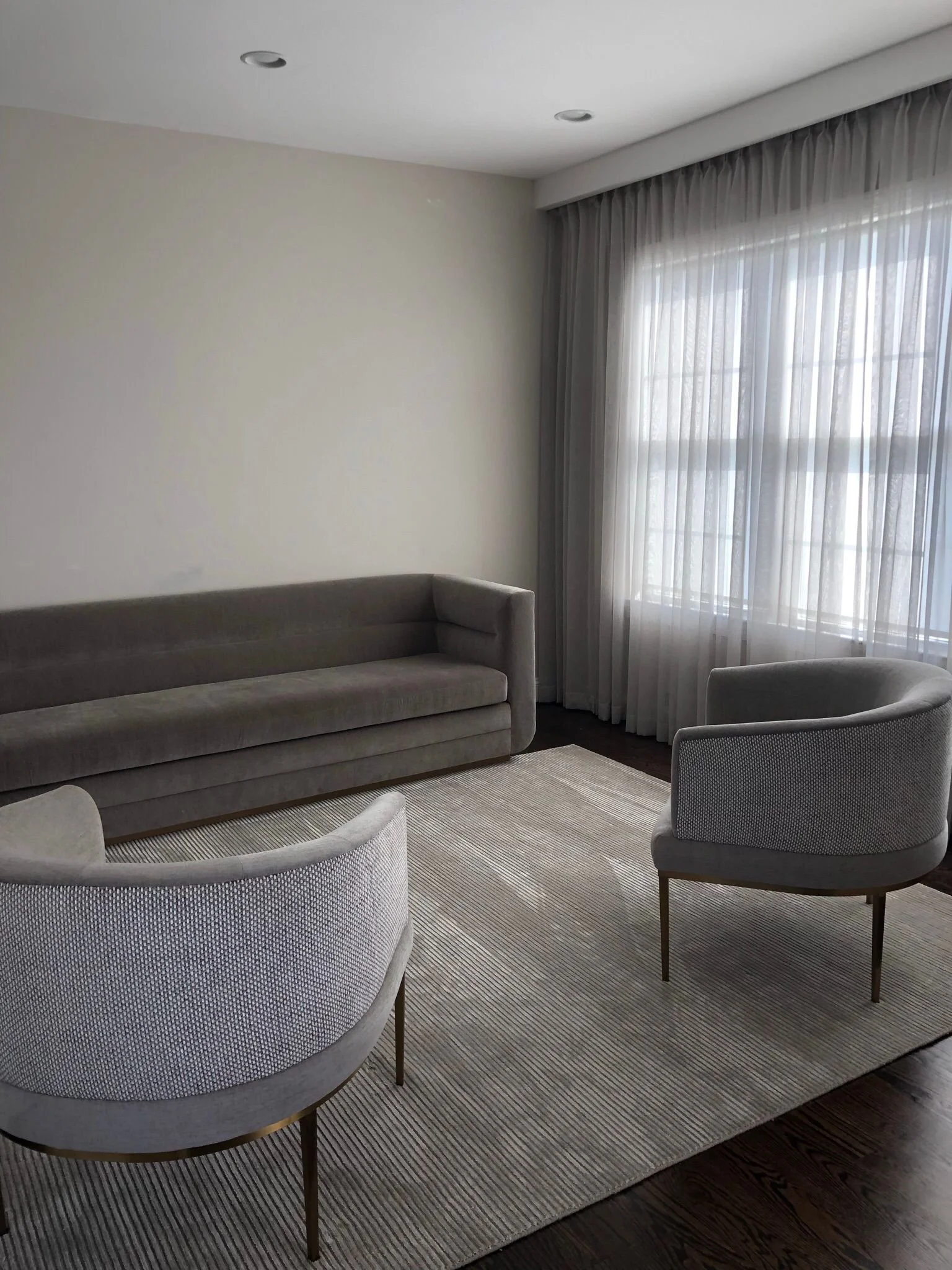 Living room with a beige sofa, two light-colored armchairs, a light-colored rug, and a large window with sheer curtains.