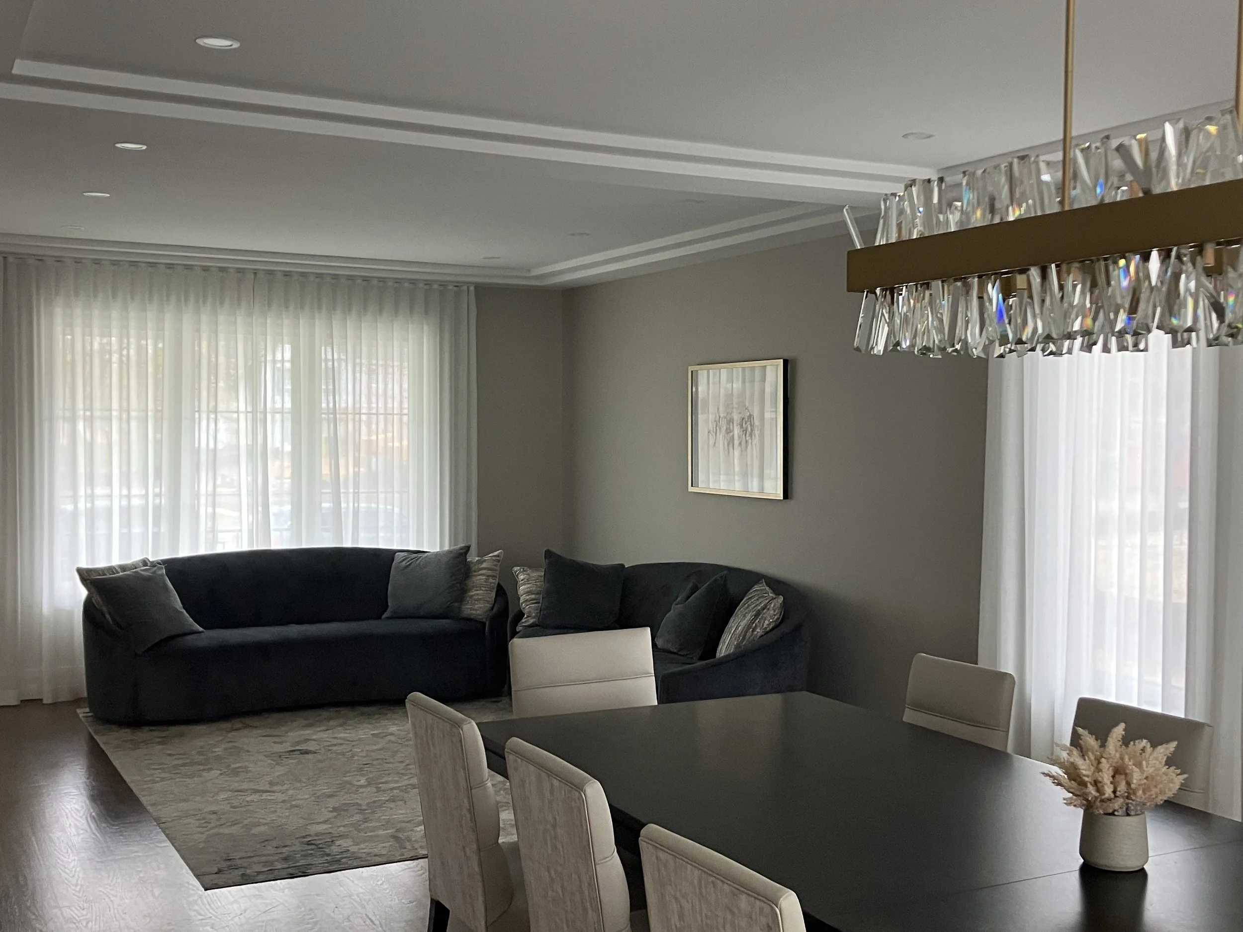 Living room with dark sofas, beige dining chairs, a large window with sheer curtains, a framed picture on the wall, a chandelier with crystal details, and a vase with dried flowers on the dining table.