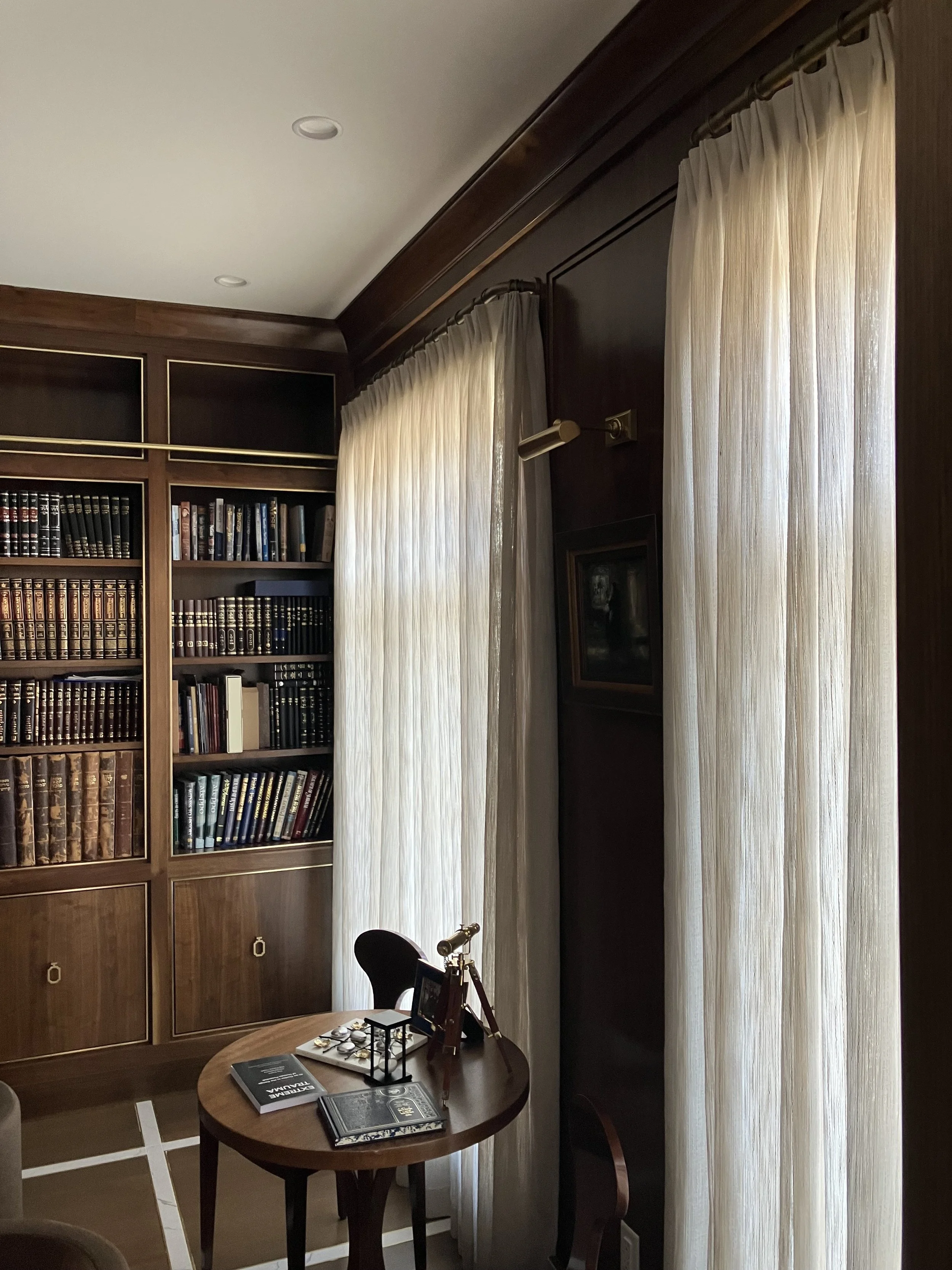 A cozy study room with wooden bookshelves filled with books, a desk with a telescope and documents, and cream-colored curtains covering large windows, illuminated by natural light.