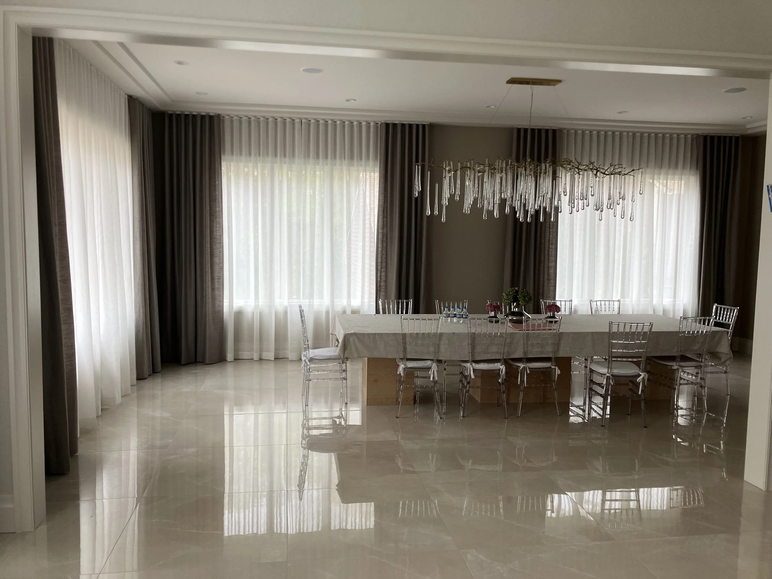 Elegant dining room with a long table, transparent chairs, a floral centerpiece, large windows with white and gray curtains, and a modern chandelier hanging above the table.