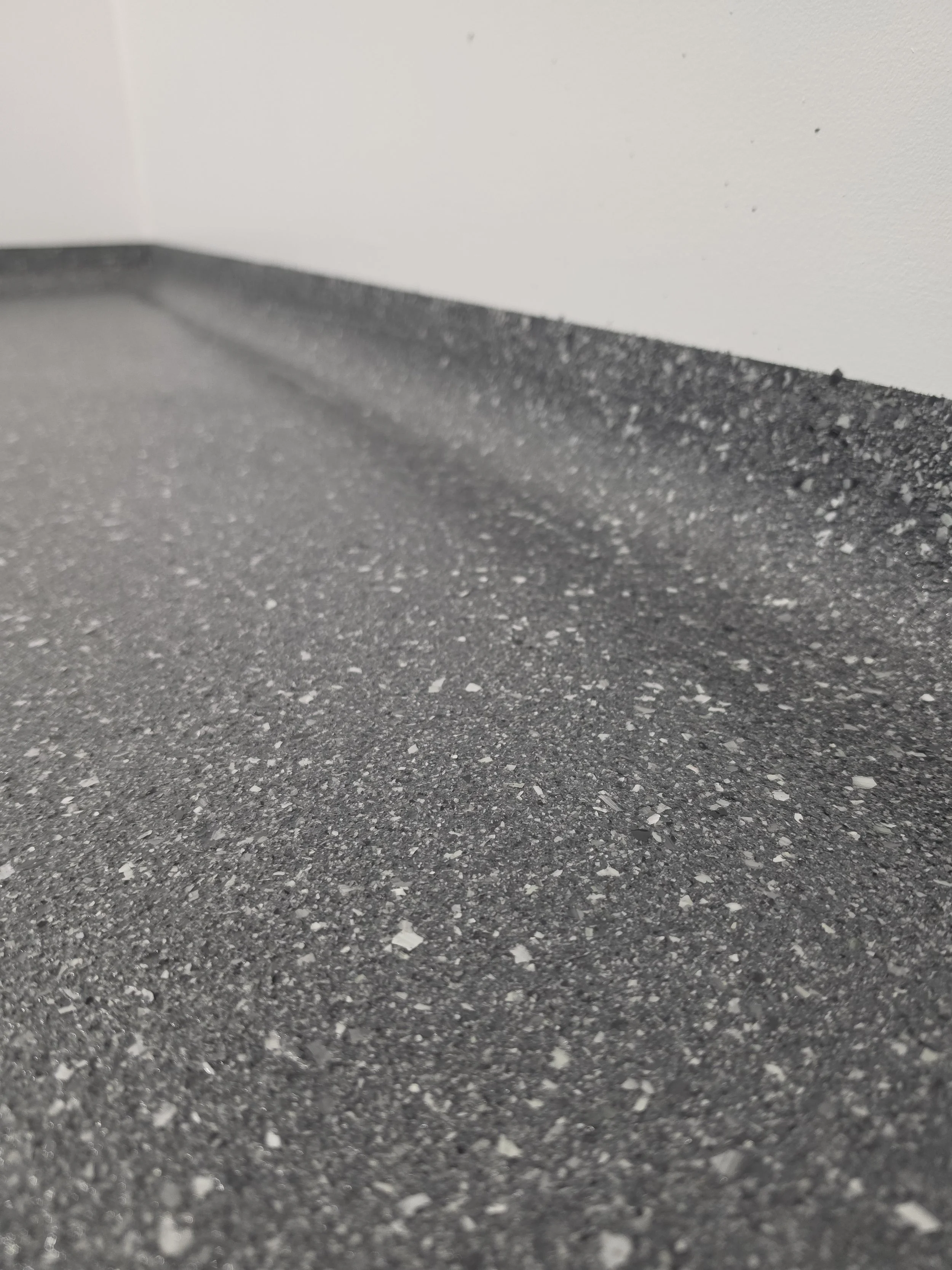 Close-up of a textured dark gray or black asphalt or concrete surface with a white wall in the background.