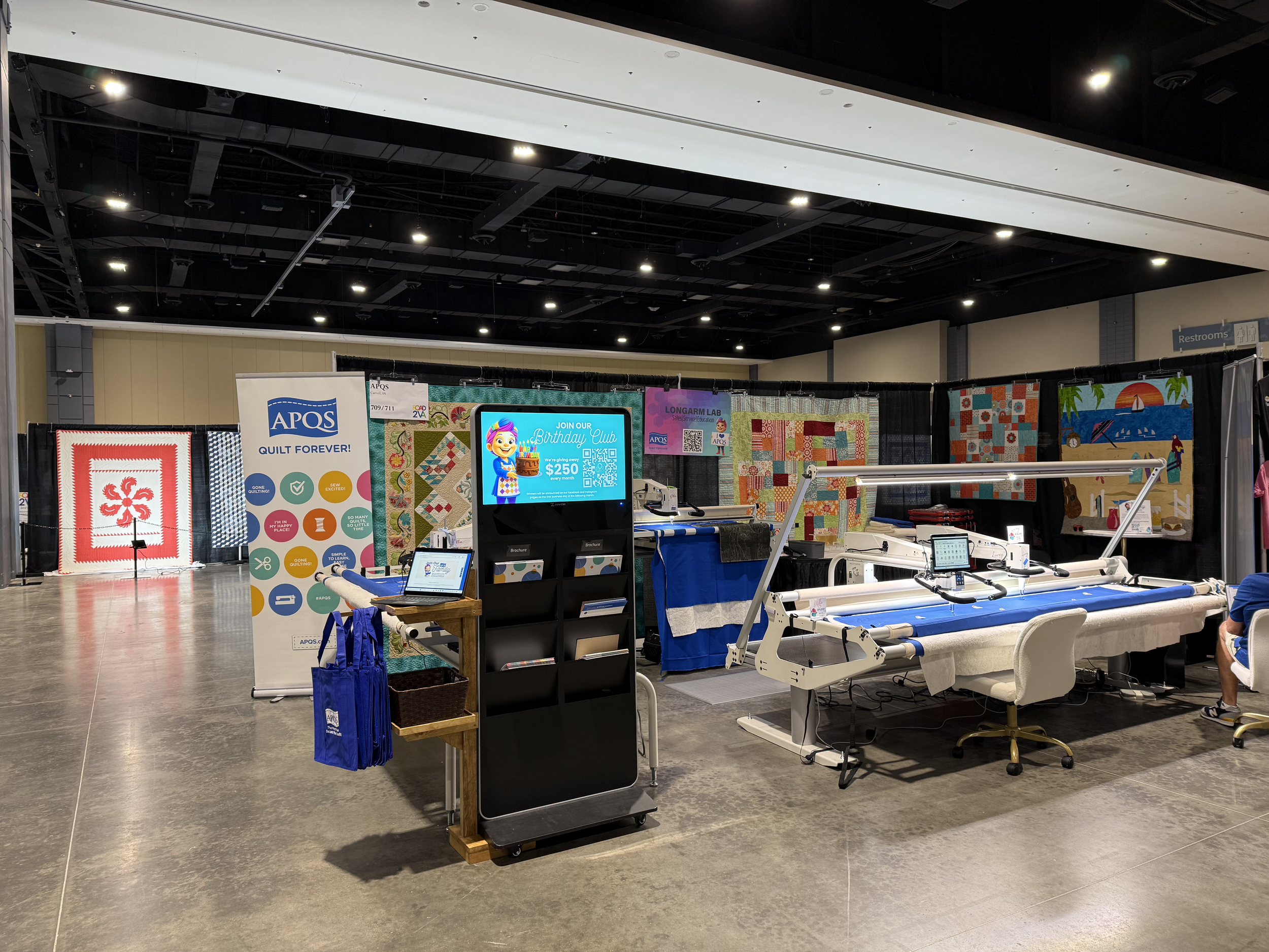 An indoor quilt booth at a convention showroom floor, with various fabric quilts on display; a digital sign promoting a birthday club through APQS, and quilting equipment including sewing machines and fabric cutting tables.