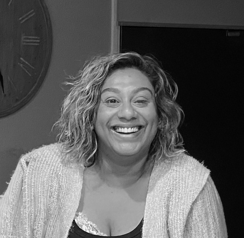 A smiling woman with curly hair sitting indoors, wearing a light-colored knitted cardigan over a black top.