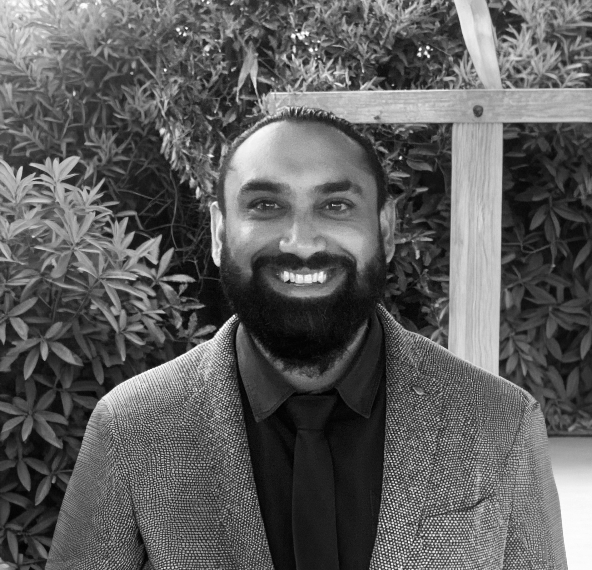 A smiling man with a beard and mustache dressed in a checked blazer, black shirt, and tie, standing outdoors in front of leafy bushes and a wooden structure.