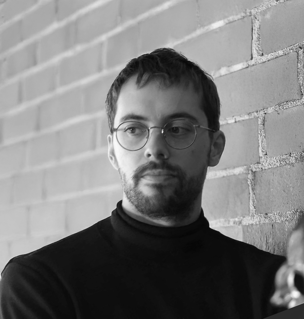 A young man with glasses and a beard, wearing a black turtleneck, standing against a brick wall, looking to his left with a serious expression.