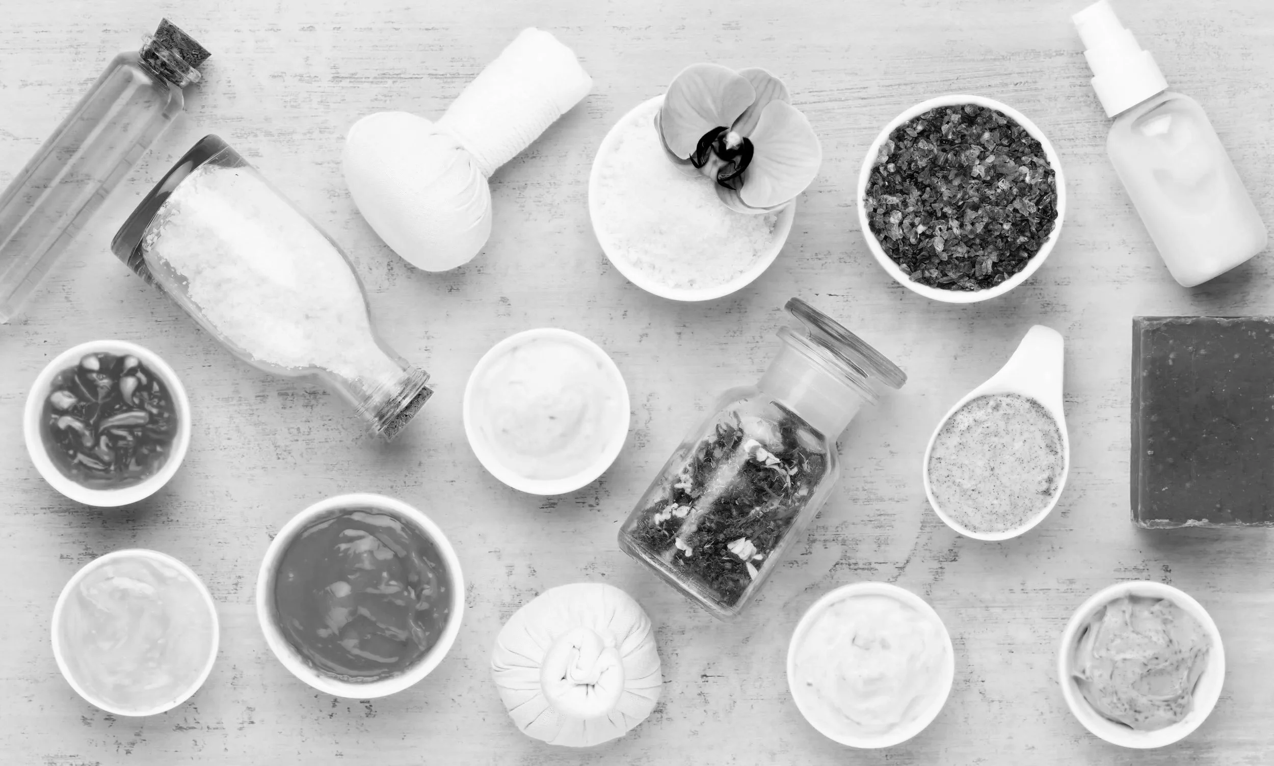 A variety of spa and skincare products arranged on a wooden surface, including jars, bottles, bowls, a soap bar, and a rolled towel.