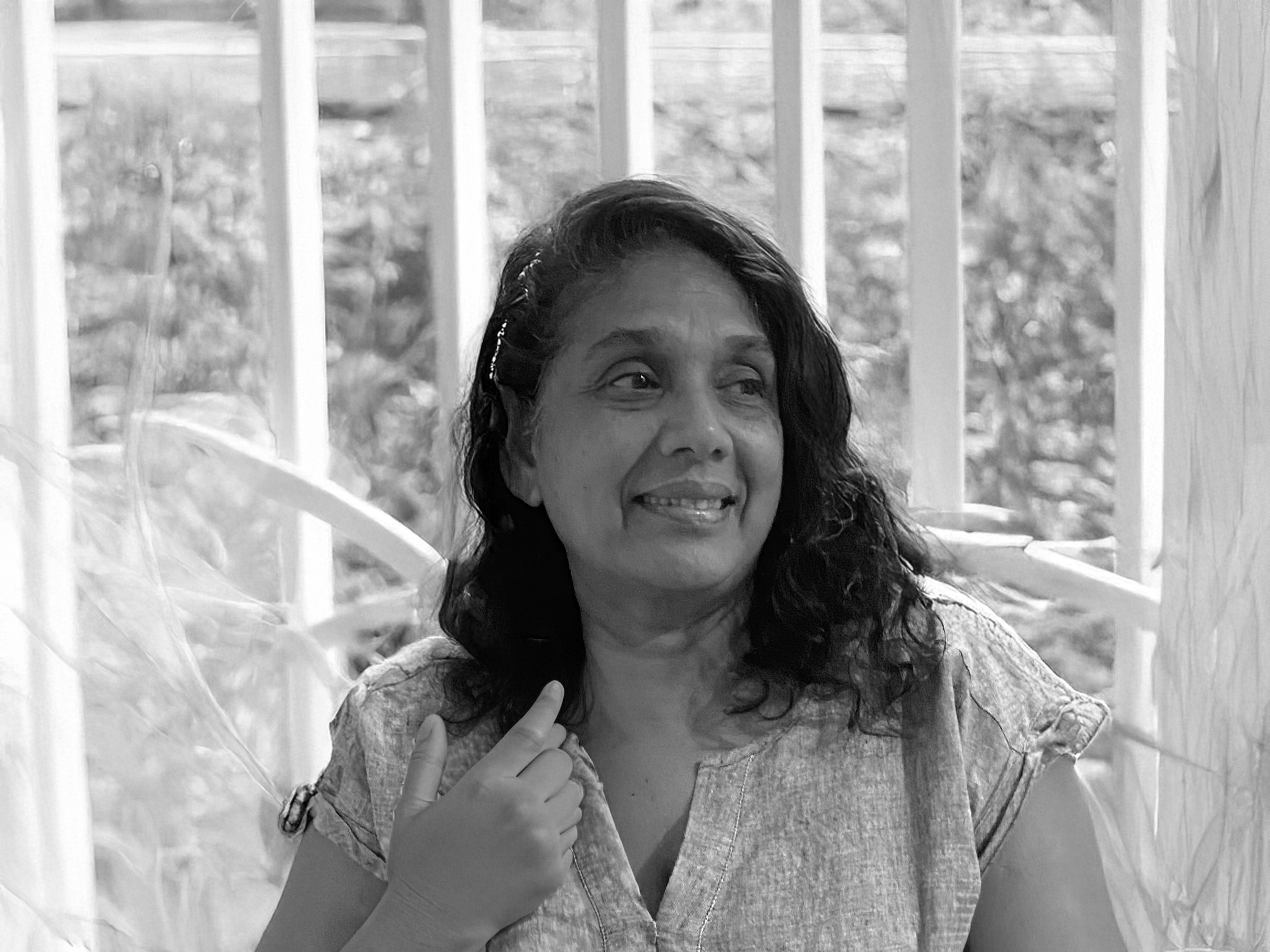 Black and white photo of a smiling woman with shoulder-length hair, wearing a sleeveless shirt, standing in front of a window with bars, with trees visible outside.