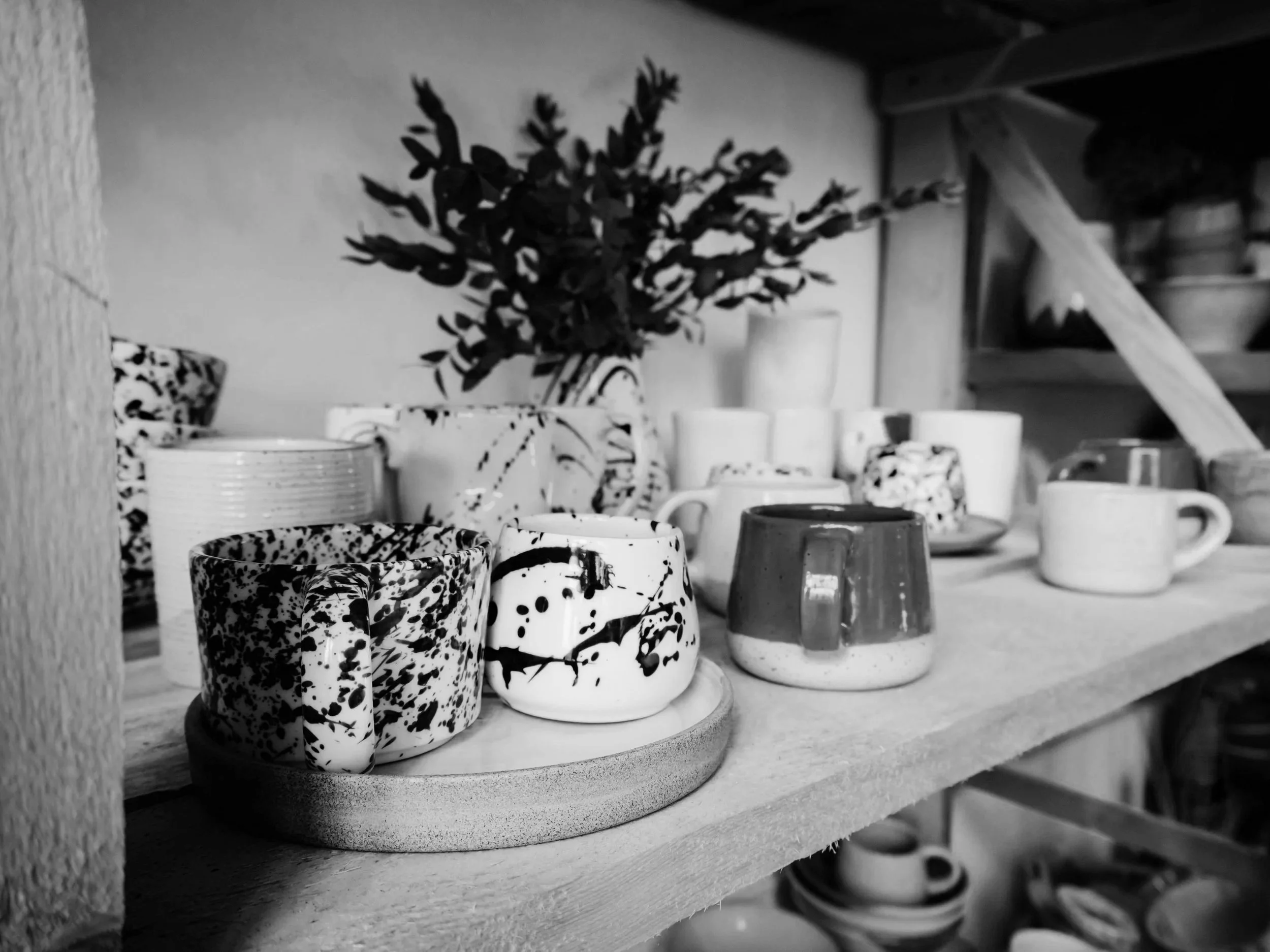 Various ceramic cups and mugs on a wooden shelf, with a vase of flowers in the background, black and white photo.