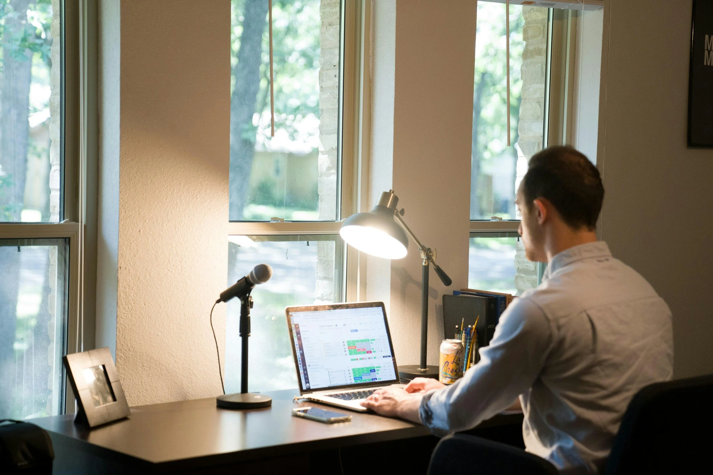 Man working on a laptop at a desk near a window, with a microphone, desk lamp, photo frame, and stationery.