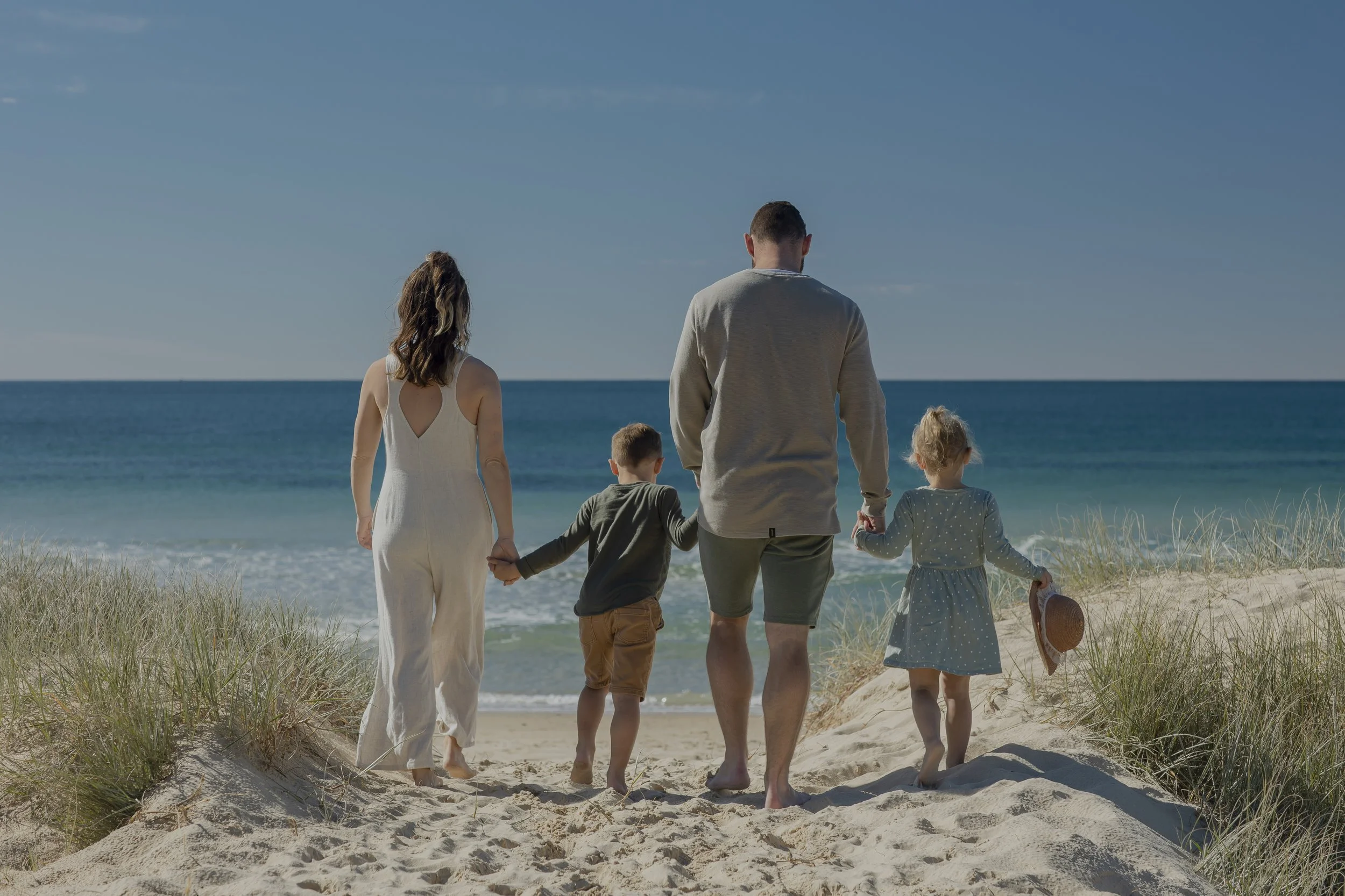 Family of four walking hand in hand on sandy beach towards ocean, with clear blue sky and grassy dunes around.