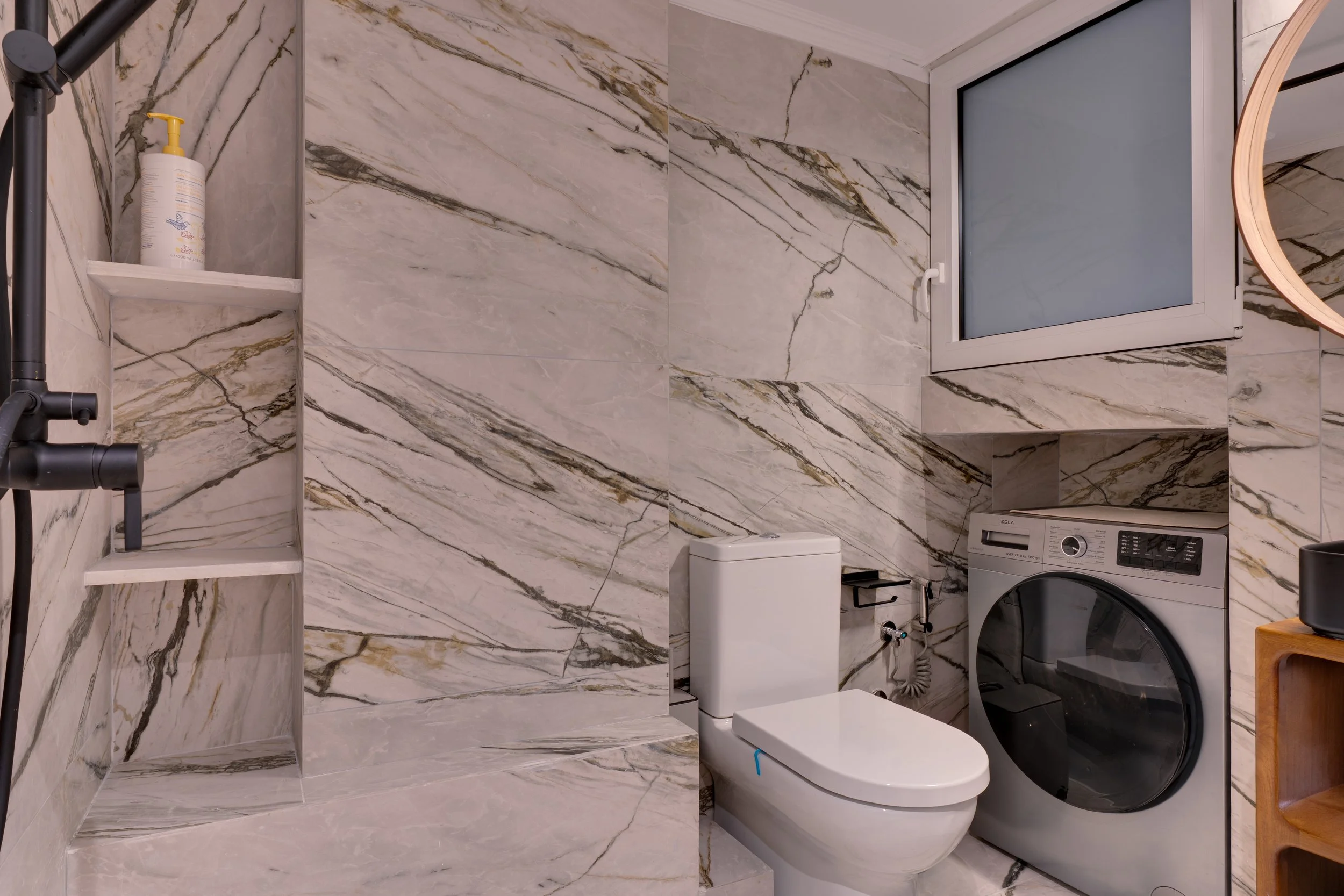Modern bathroom with marble walls and floor, small window, black shower head and shelf, washing machine, toilet, small white cabinet, and wooden shelf.