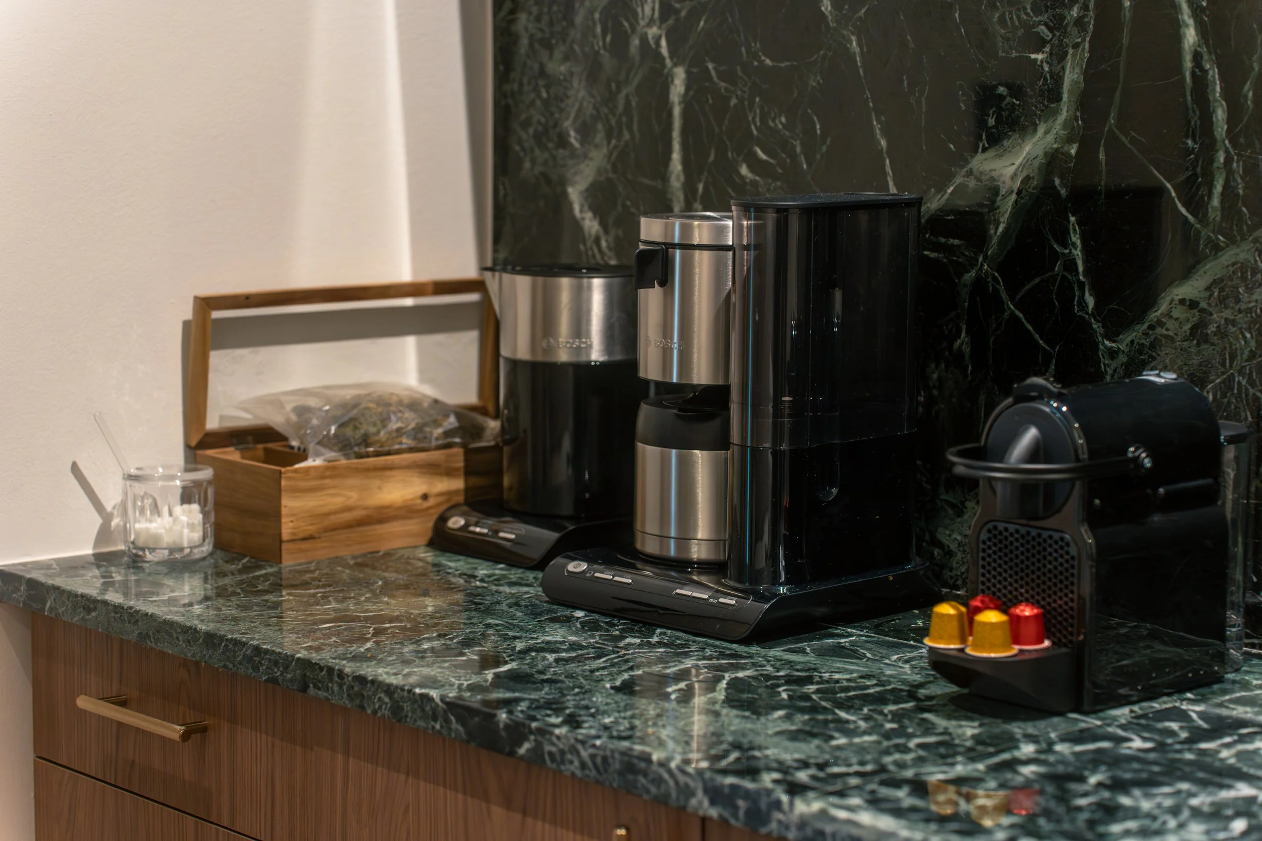 Keurig coffee maker with a stainless steel mug on a black tray on a green marble countertop. To the left, a glass jar with sugar, and a wooden box with tea bags. Small colorful capsules are on a black tray in the foreground. Background features a bla