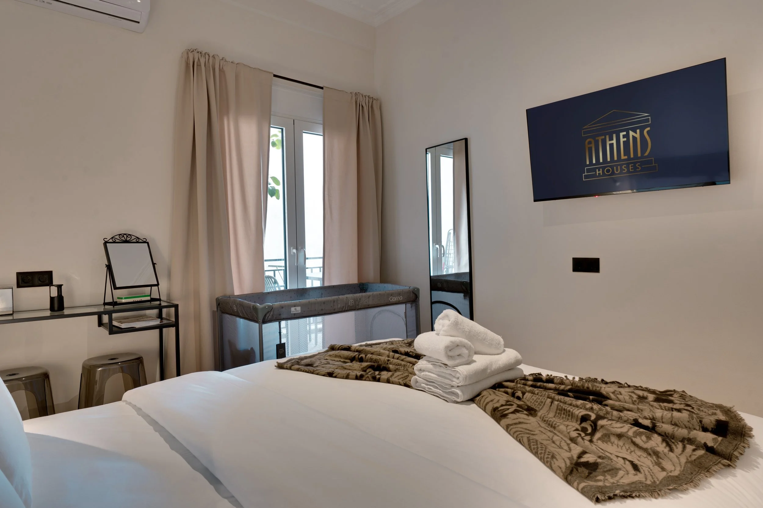Hotel room with two beds, a TV on the wall displaying 'Athens Houses,' a sliding glass door with cream curtains, a mirror, a small table, and a baby crib. Towels are placed on the bed.