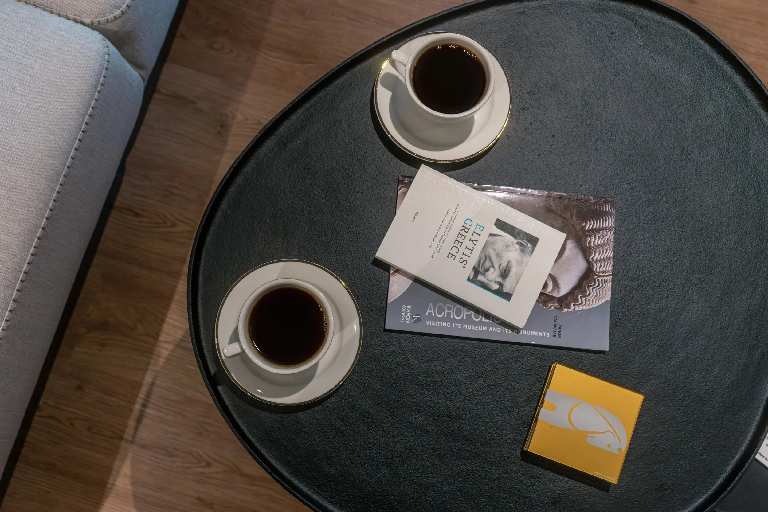 Two cups of black coffee on saucers on a dark round table. A magazine with the title 'ACROPOLIS' and a brochure titled 'Elvytis Greece' are also on the table. The background includes part of a light-colored couch and a wooden floor.