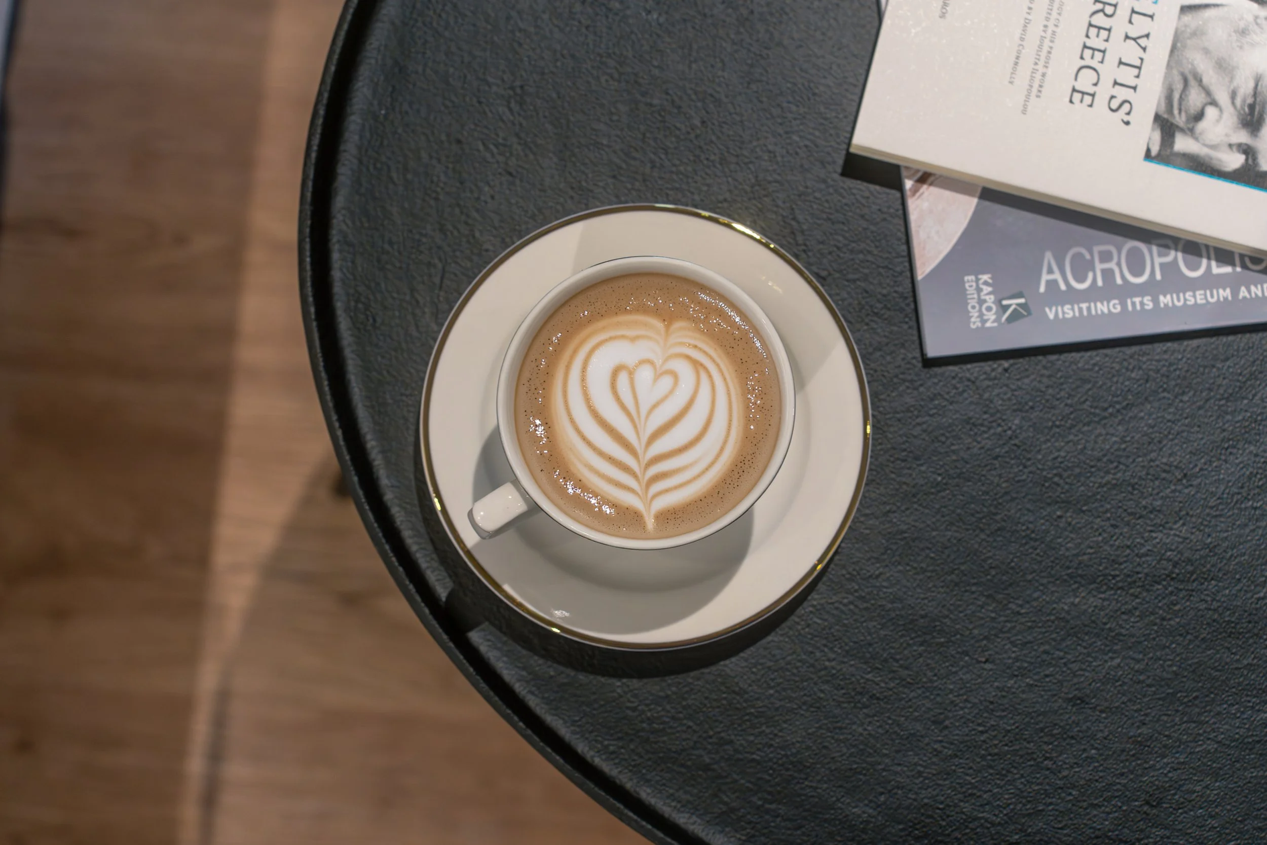 A cup of coffee with heart-shaped latte art on a black table next to a magazine and a book.