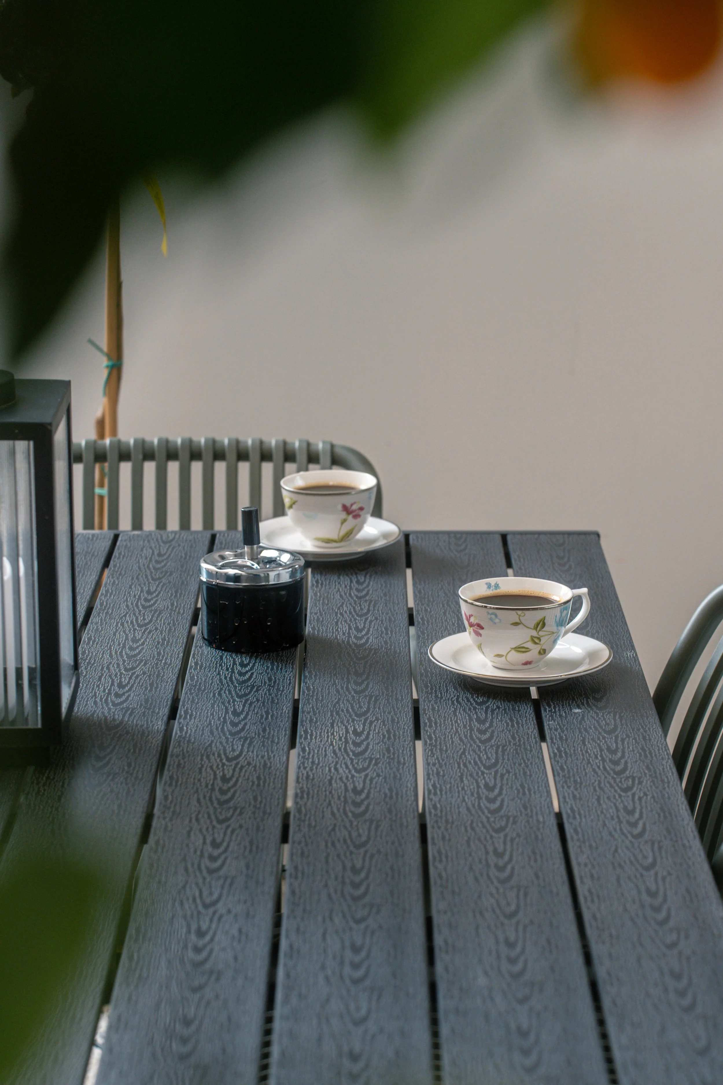A table with two cups of coffee, a black thermos, and part of a lantern, with greenery in the foreground and a plain wall in the background.
