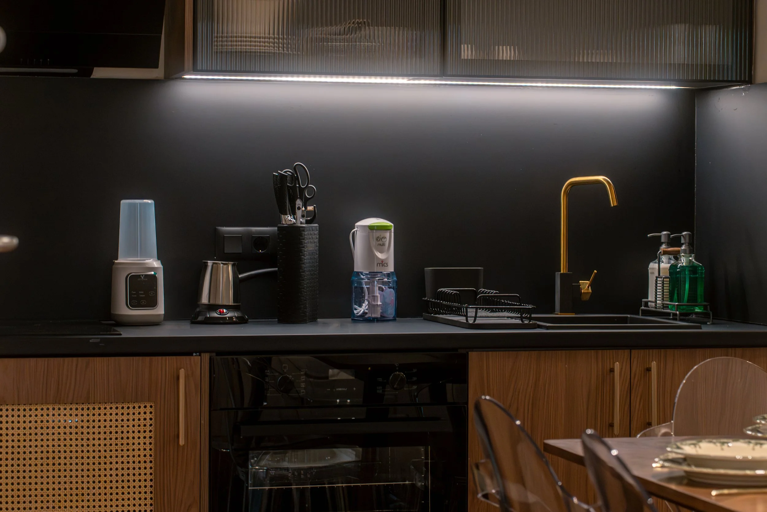A modern kitchen with a dark countertop, wooden cabinets, and various small appliances, including a blender, kettle, and a bottle of hand soap near a gold-colored faucet.