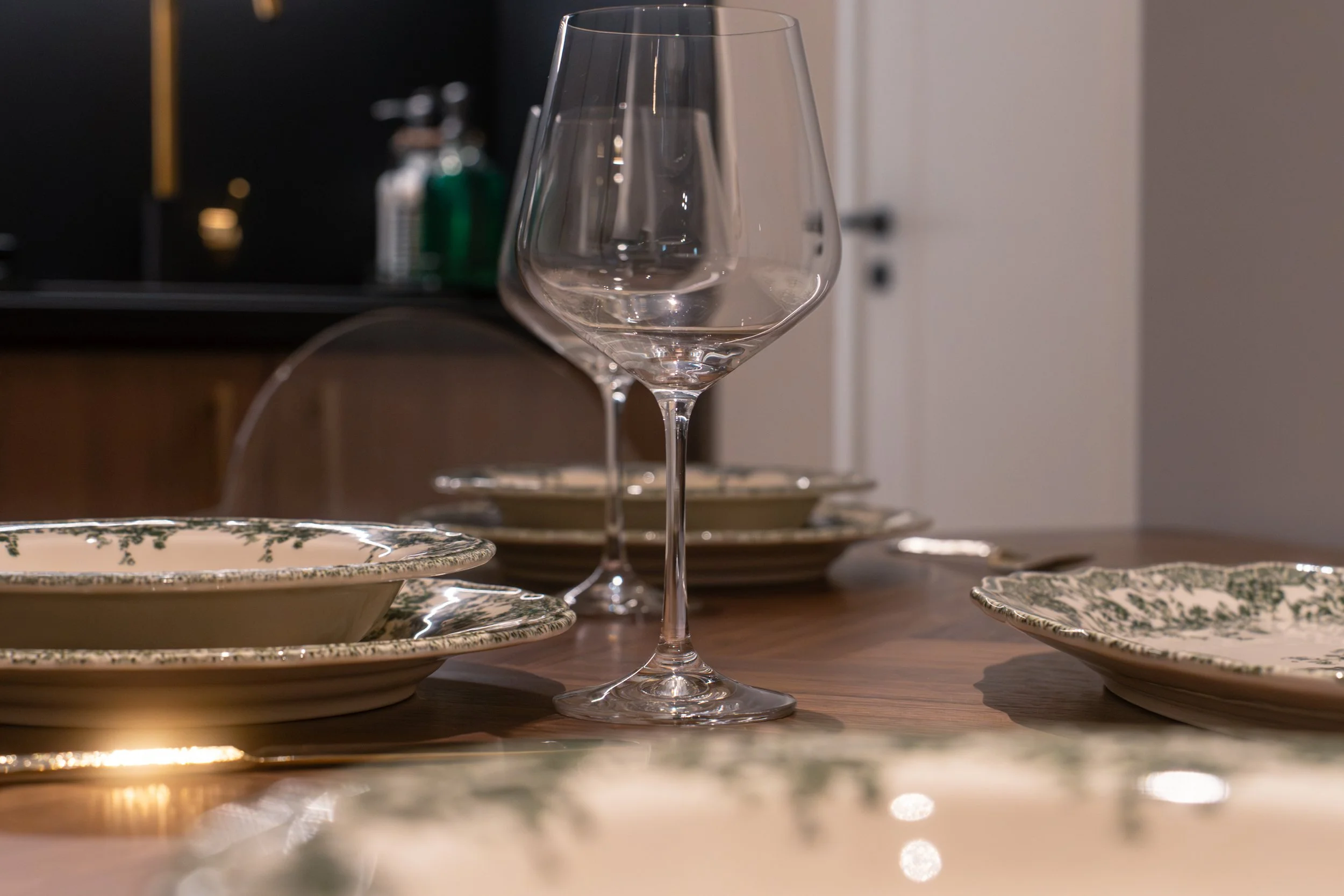 A partially filled wine glass on a wooden table surrounded by floral-patterned dishes, with a blurry background including a bottle and a black object.