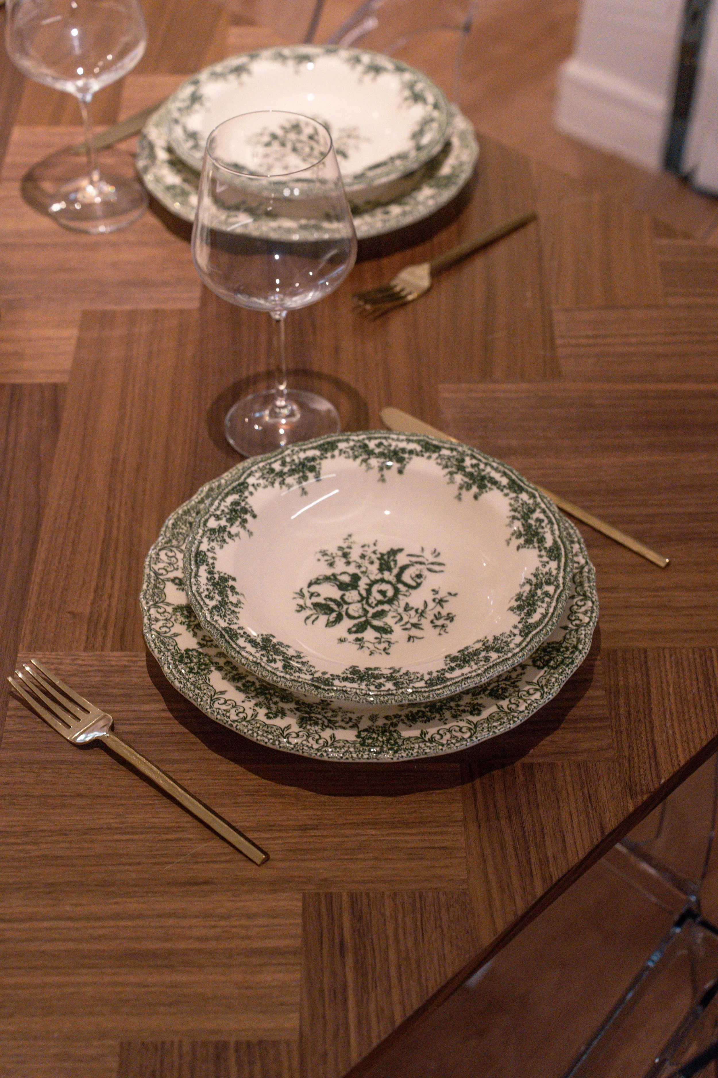 A dining table set with patterned china plates, clear water glasses, gold-colored forks, and a wooden table.