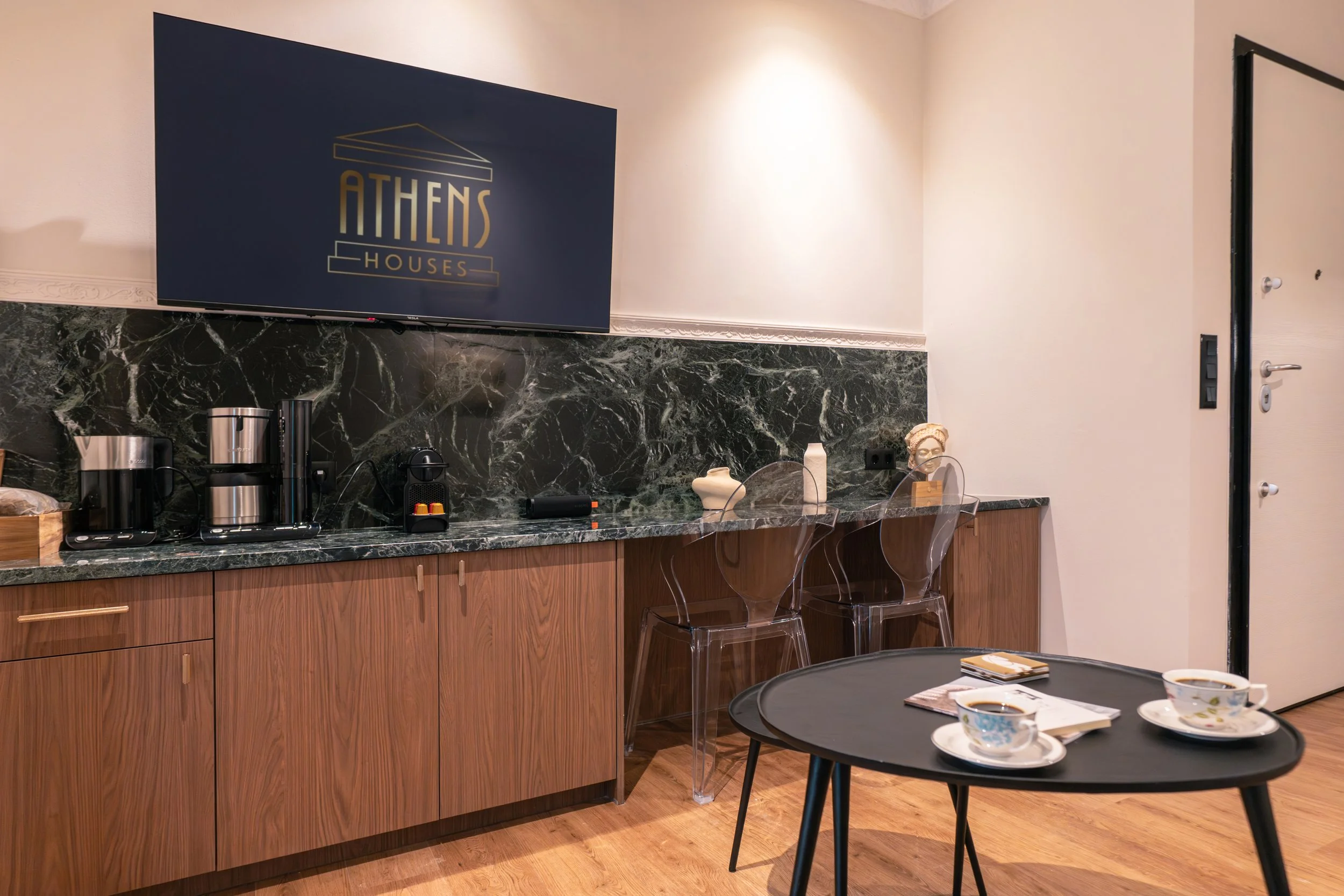 A modern kitchen or break room with a dark marble countertop, wooden cabinets, and a wall-mounted TV displaying a logo that says 'Athens Houses.' On the counter are coffee machines and a coffee maker. There are three clear acrylic chairs at the count