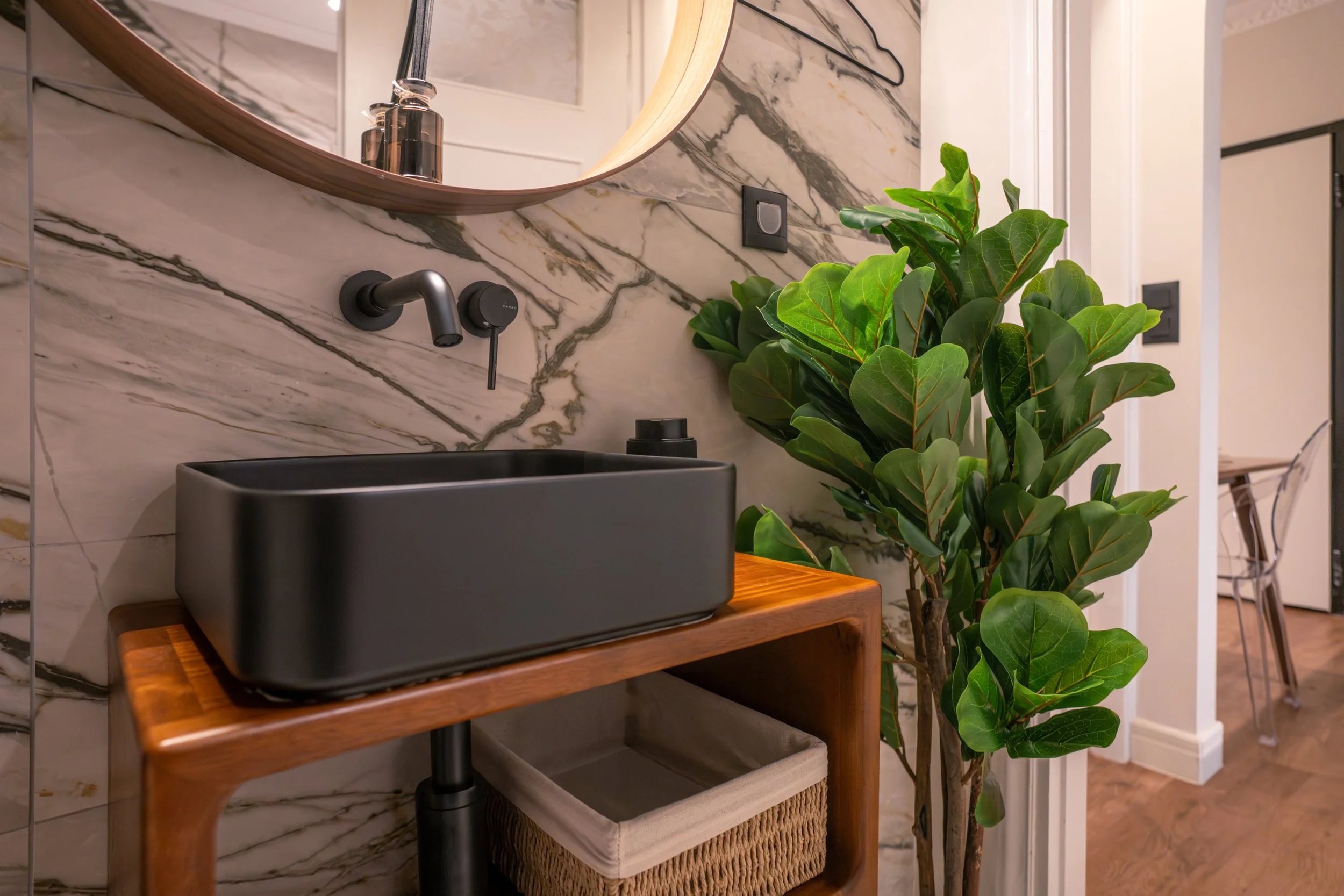 Modern bathroom with marble wall, black sink on wooden stand, large green leafy plant, mirror, and black faucet.