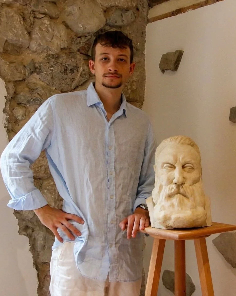 Un homme debout à côté d'une sculpture de tête en pierre d'un homme avec une barbe et une moustache, dans une pièce avec un mur en pierre et un mur blanc.