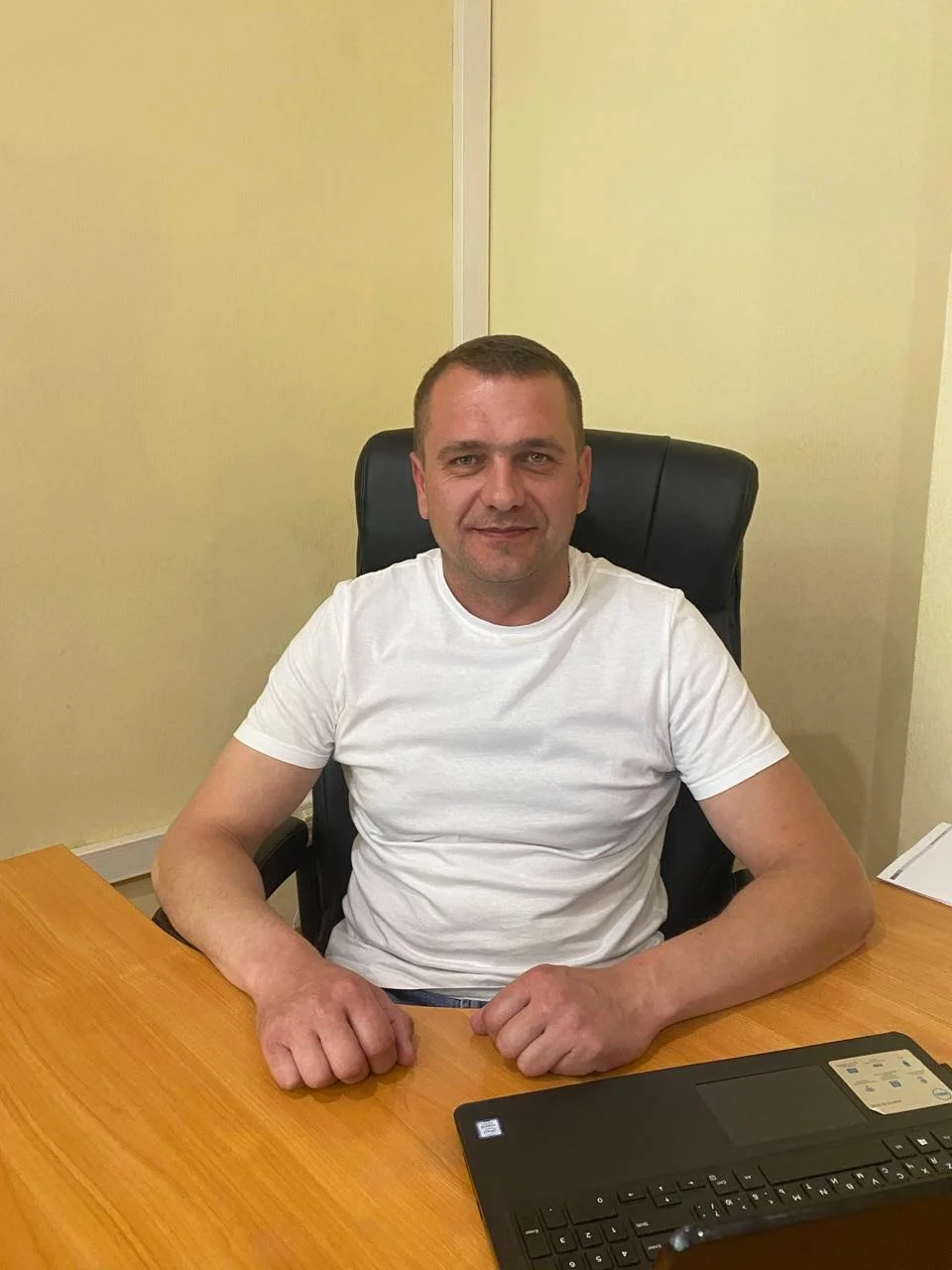 A man sitting at a wooden desk in an office, wearing a white t-shirt, with a black office chair and a laptop in front of him.