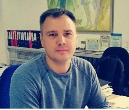 A man with short dark hair wearing a gray long-sleeve shirt sitting at a desk in an office, with binders and framed documents on the wall behind him.
