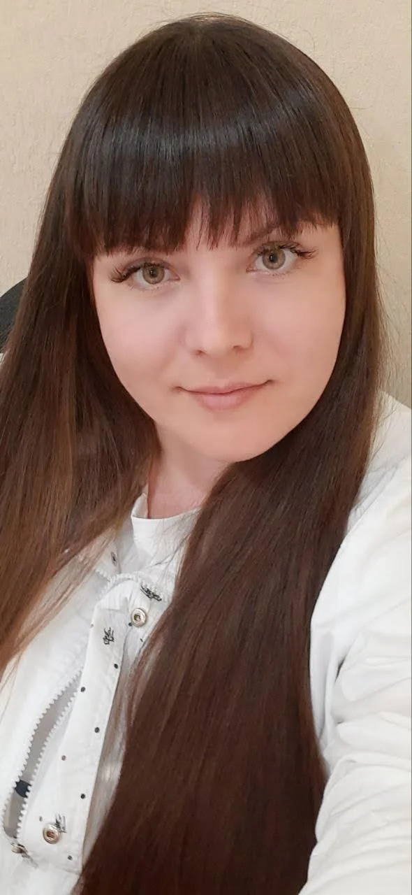 A woman with long, straight, brown hair and bangs, wearing a white top, smiling softly at the camera inside a room with beige walls.