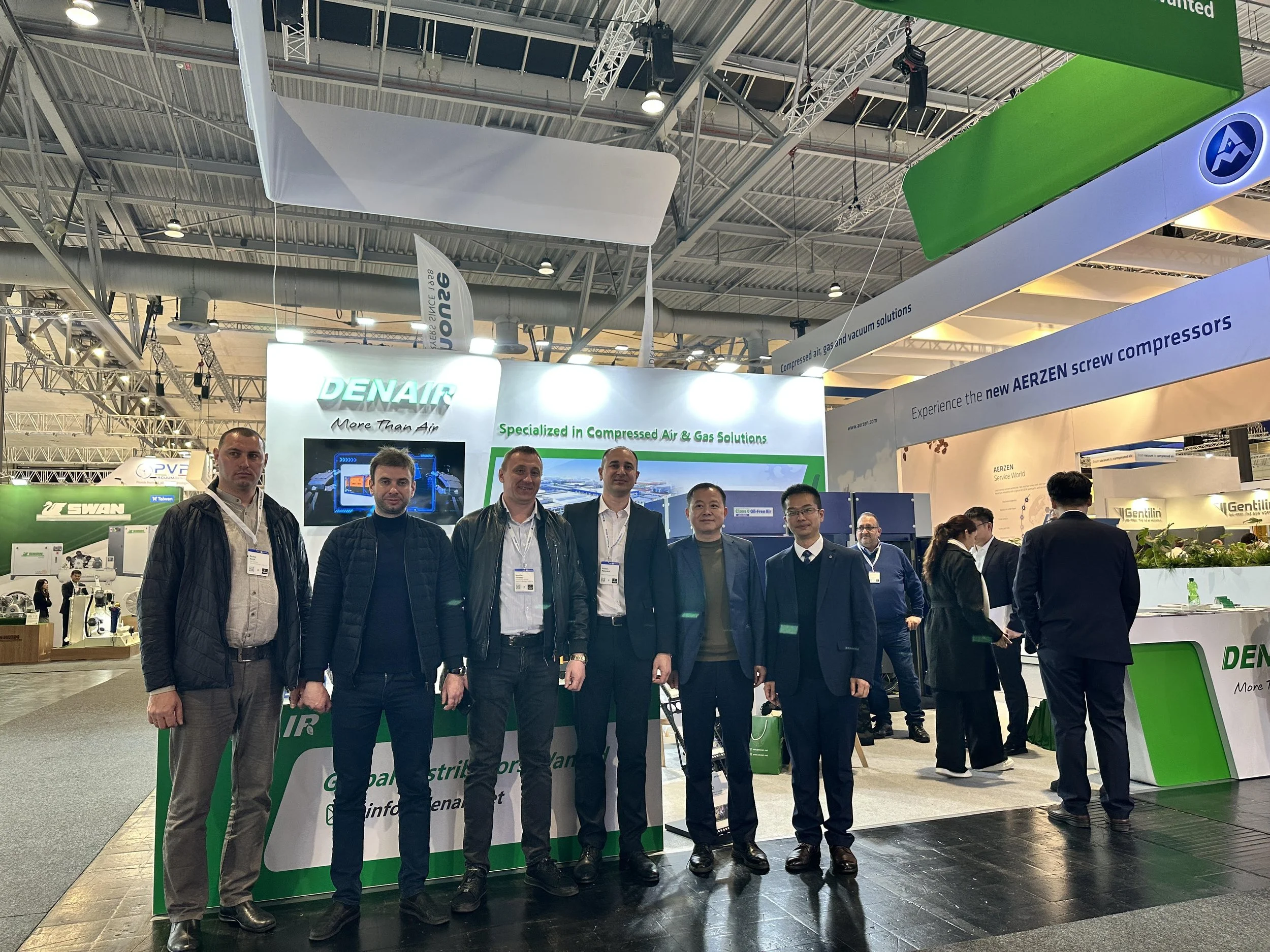 Group of seven men standing in front of a DENAIR booth at an indoor exhibition, with banners and displays related to compressed air and gas solutions.