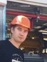 A young man wearing an orange hard hat standing in an industrial or construction setting.