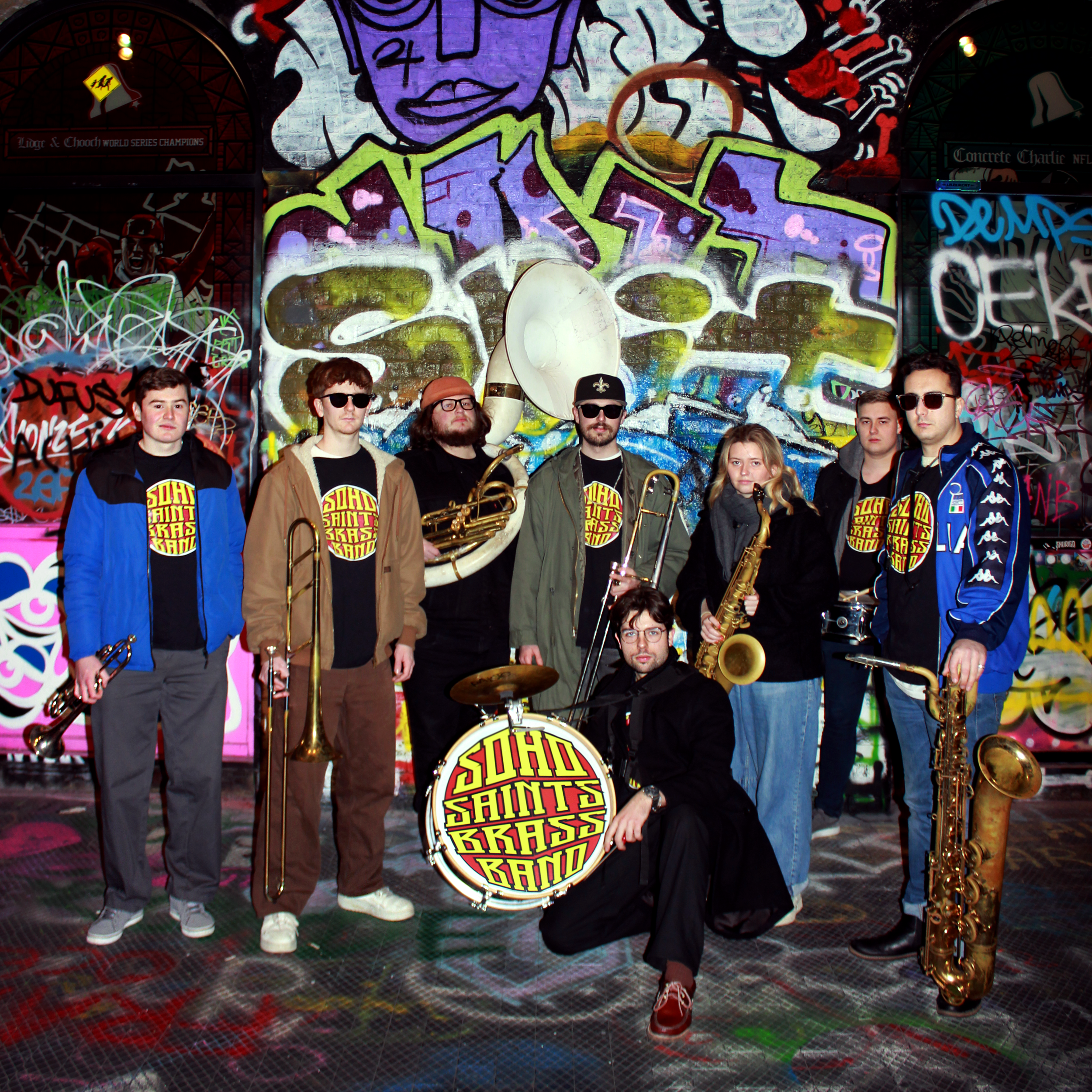 A band called 'Soho Saints Brass Band' posing in a colorful, graffiti-covered alleyway with musical instruments. The band members are holding various brass instruments, and they are standing in front of a large, vibrant mural.
