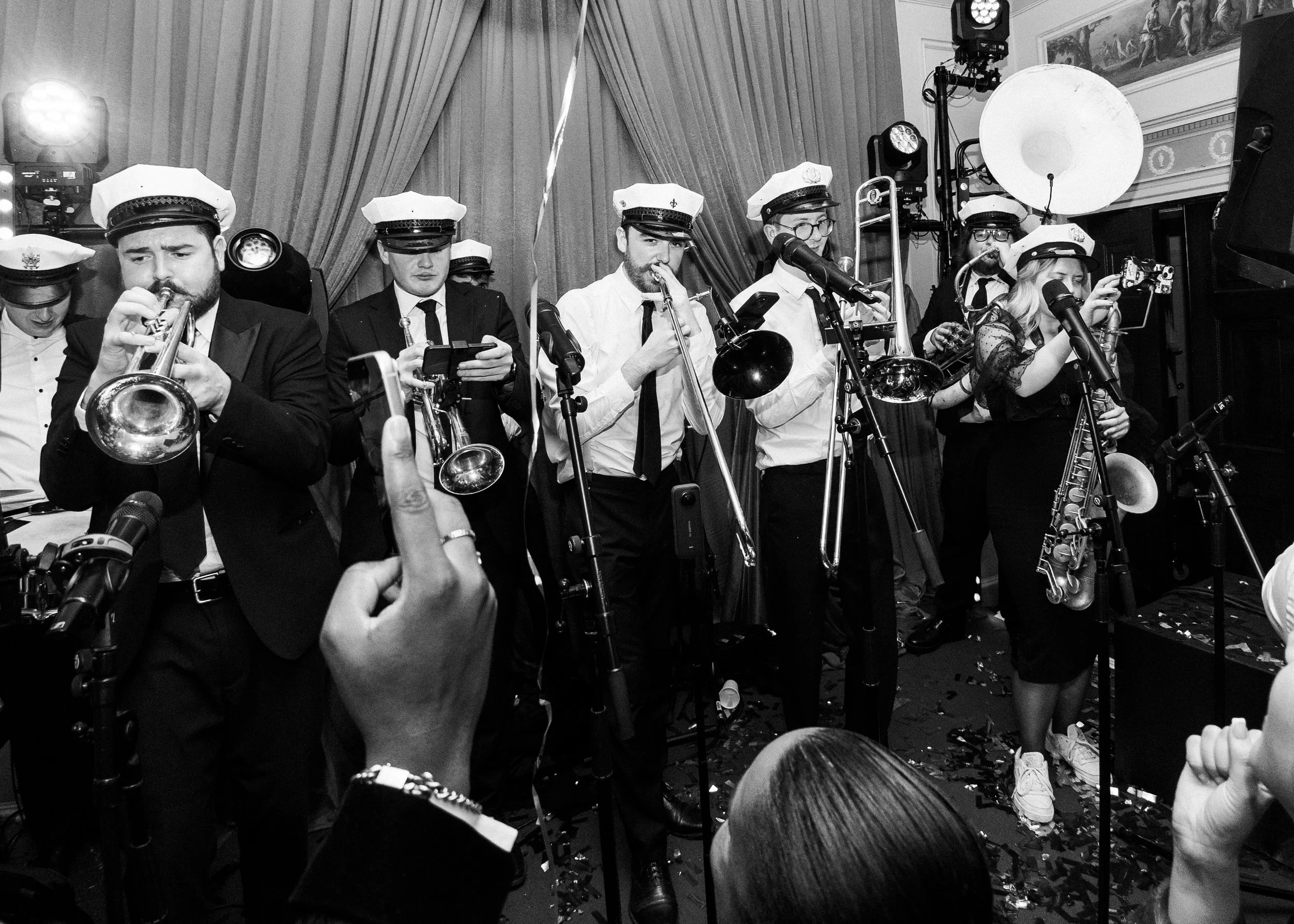 A black and white photo of a band performing at a celebration event, all members wearing white hats, with one person in the audience taking a photo.