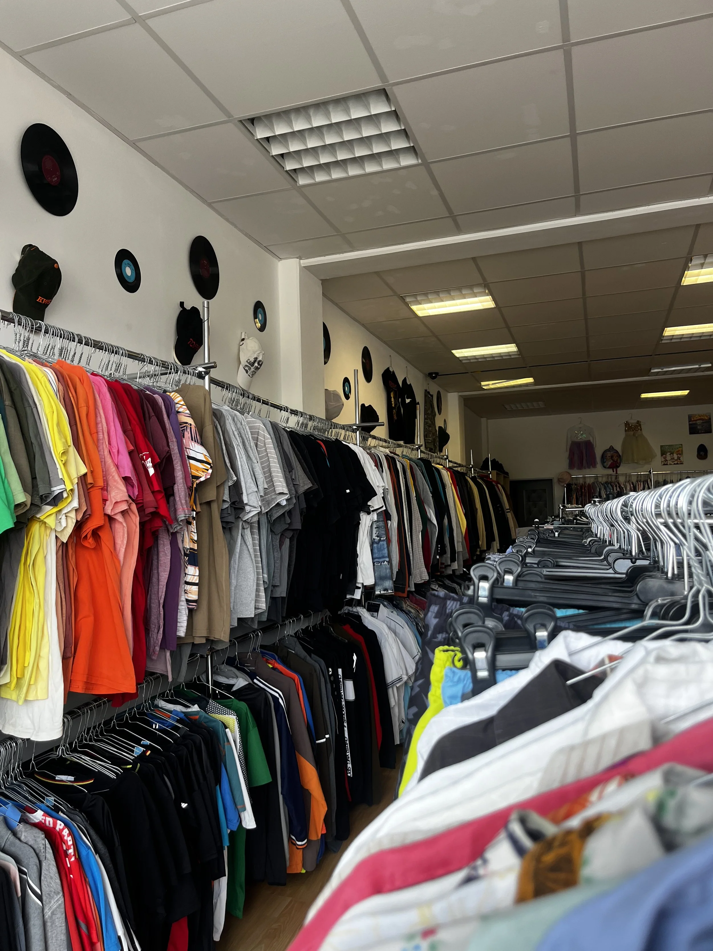 Sara Second Hand in Eisenhüttenstadt, Second Hand & Vintage Mode für alle. Kleidung, Haushaltsartikel & Accessories. Kleiderladen mit Farbenfrohen Hemden und T-Shirts auf Haken und Regalen, Wanddekorationen mit Schallplatten und Hüten.