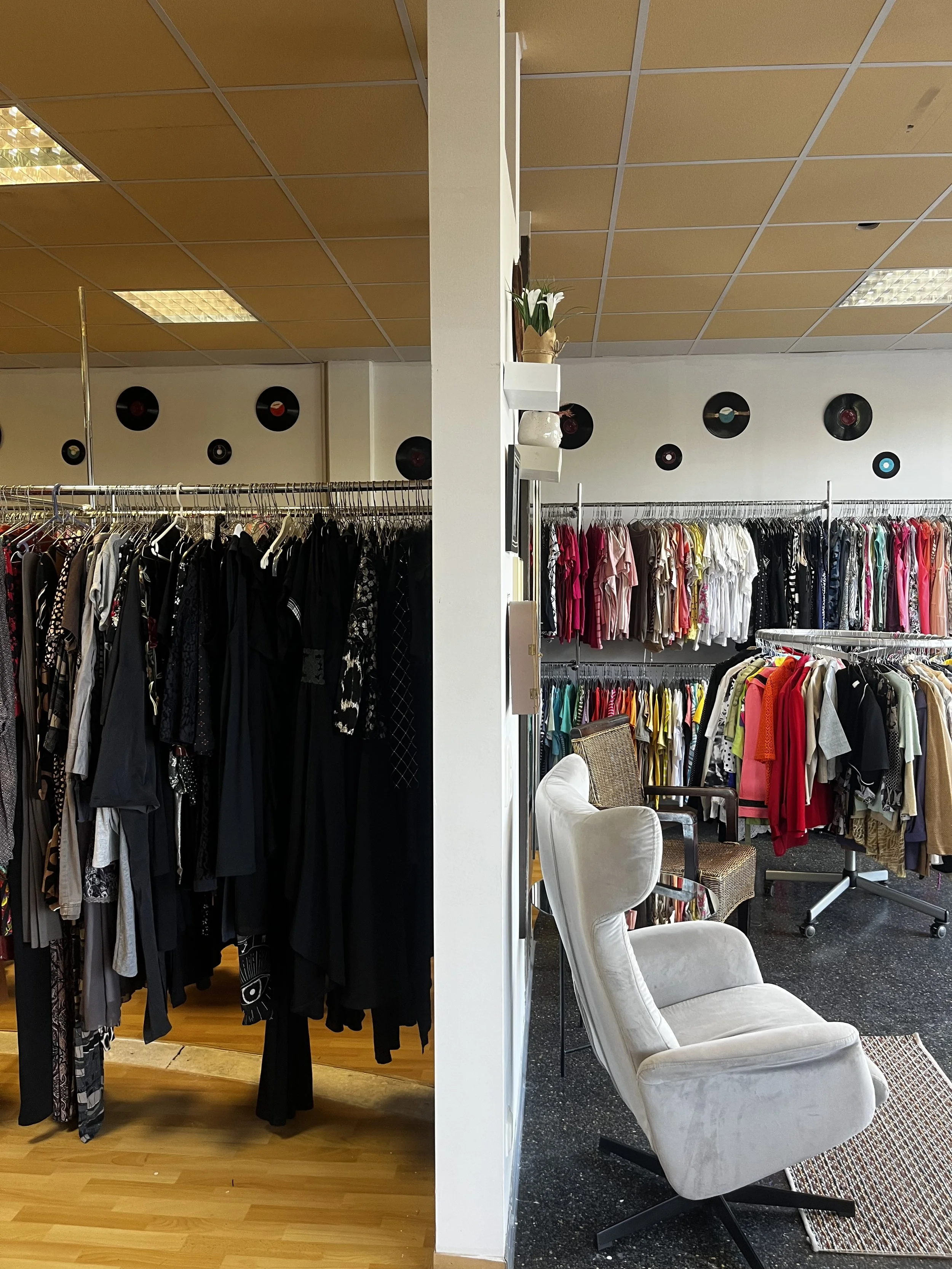 Sara Second Hand in Eisenhüttenstadt, Second Hand & Vintage Mode für alle. Zwei Raumabschnitte eines Kleidergeschäfts, getrennt durch eine weiße Wand. Links befinden sich Kleider auf Kleiderstangen, rechts eine bequemer Sitzecke zum ausruhen.