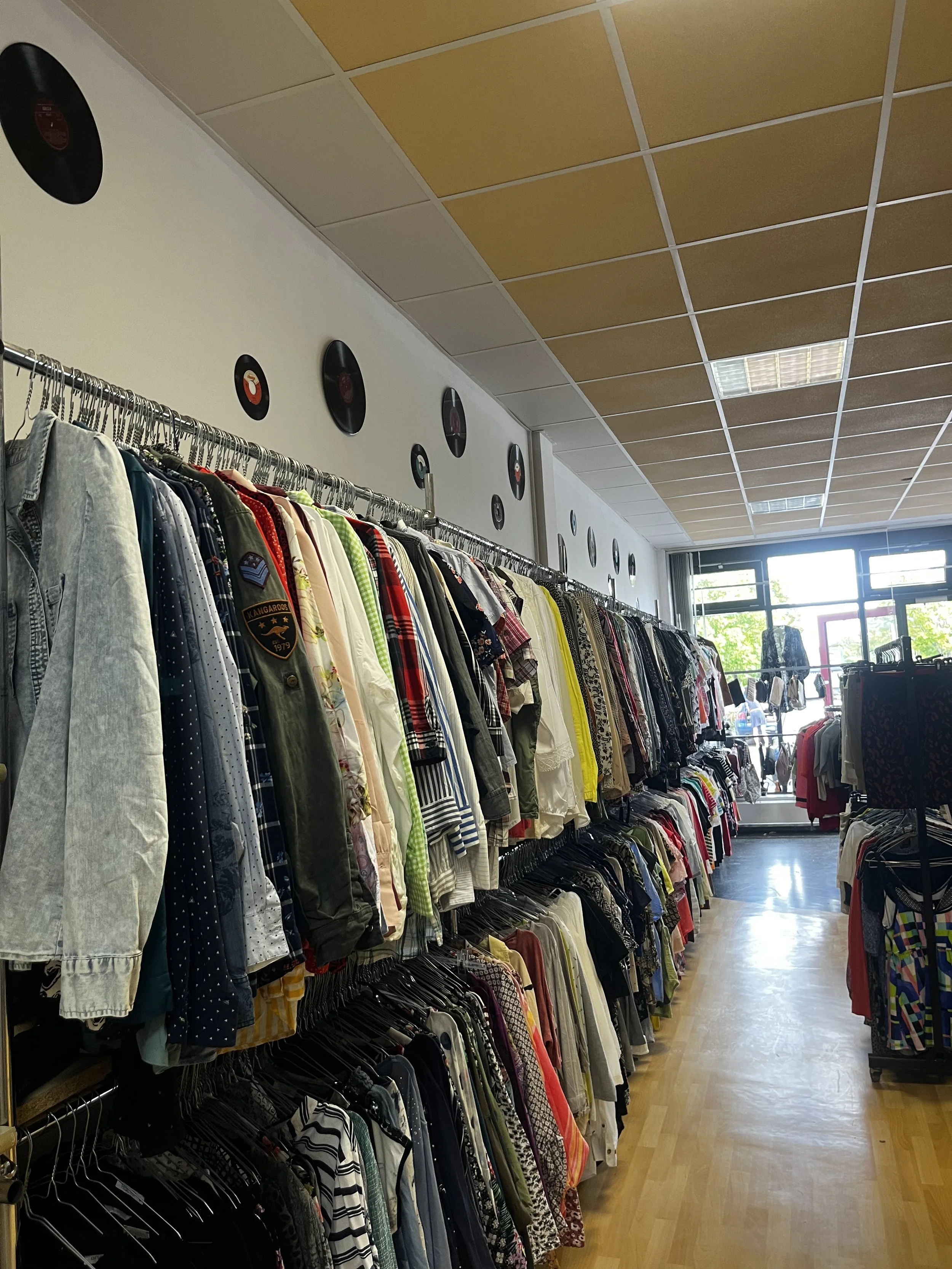 Sara Second Hand in Eisenhüttenstadt, Second Hand & Vintage Mode für alle. Kleidung, Haushaltsartikel & Accessories. Secondhandladen mit Kleidungsstücken auf Kleiderstangen und Vinyl-Schallplatten an der Wand.