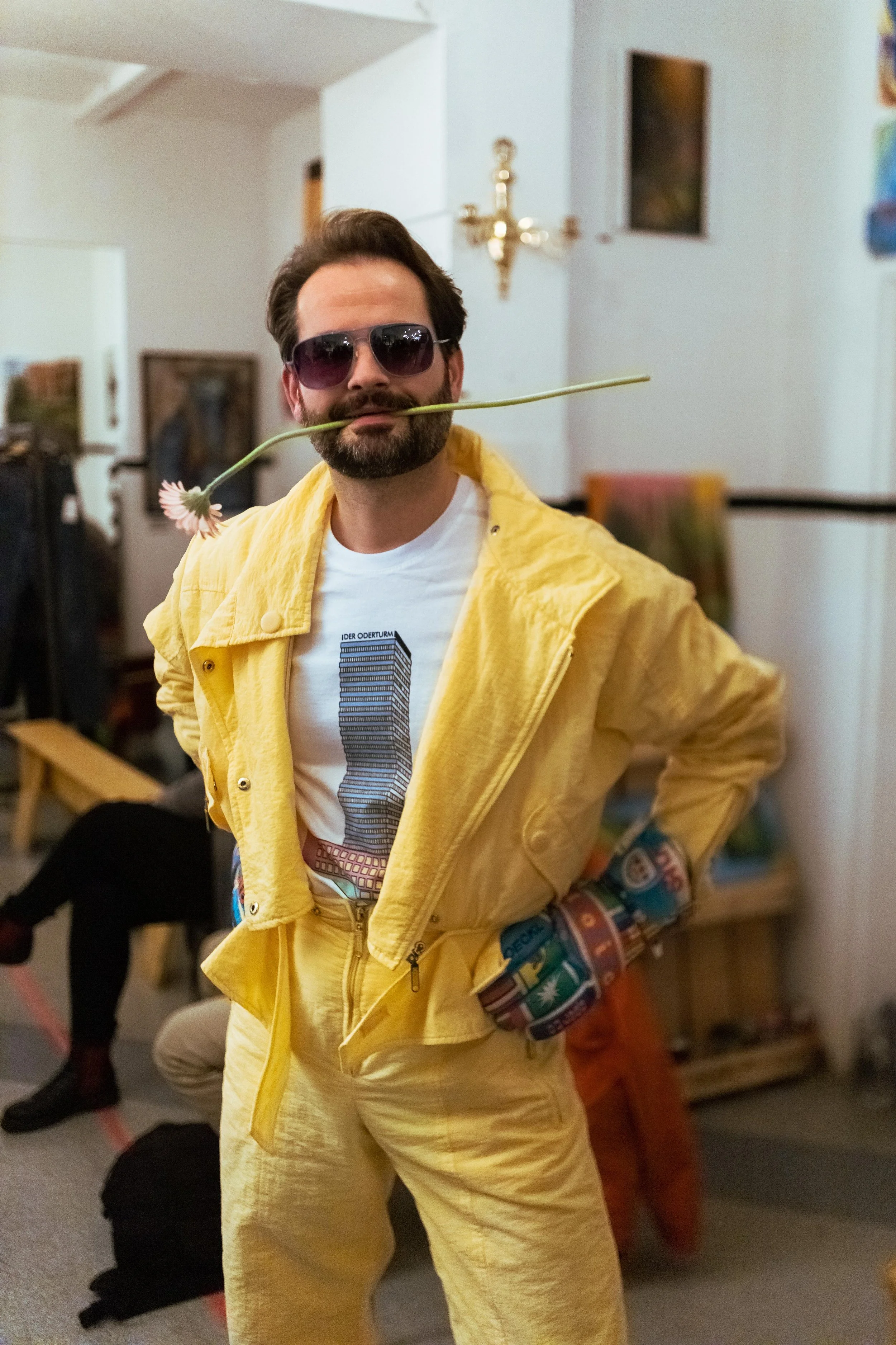 Sara Second Hand in Eisenhüttenstadt, Second Hand & Vintage Mode für alle. Kleidung, Haushaltsartikel & Accessories. Ein Model trägt einen Vintage Overroll und Sonnenbrille und hält eine Blumenstängel im Mund, während er bei einer Party ist.