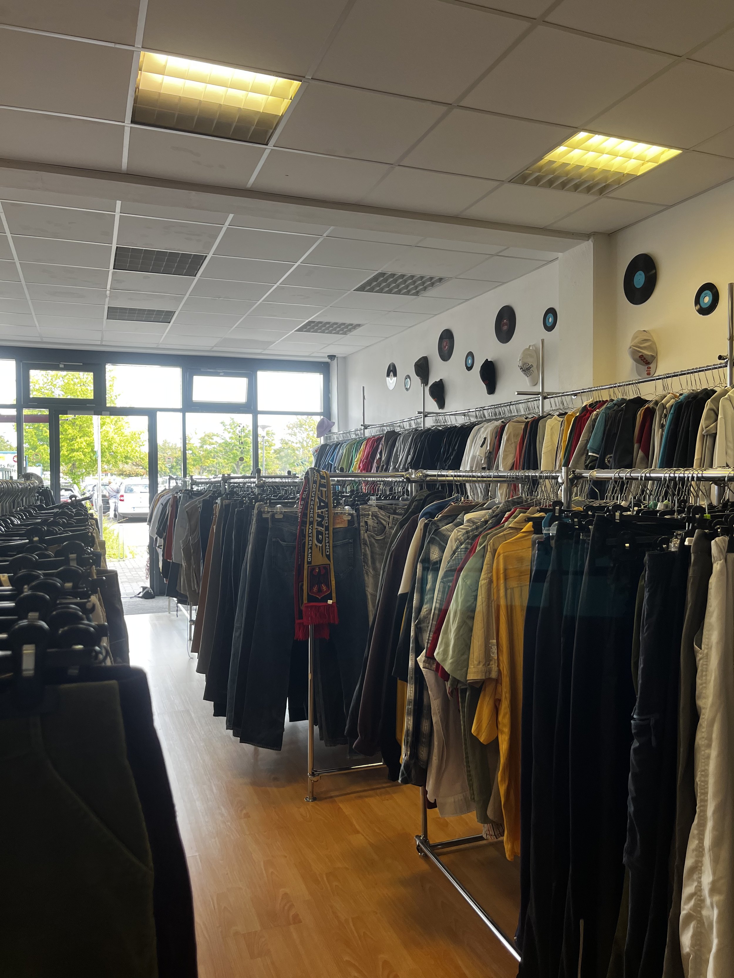 Sara Second Hand in Eisenhüttenstadt, Second Hand & Vintage Mode für alle. Kleidung, Haushaltsartikel & Accessories. Innenansicht der Kleidung auf Kleiderstangen und Vinyl-Schallplatten an der Wand, Sonnenlicht durch große Fenster im Hintergrund.