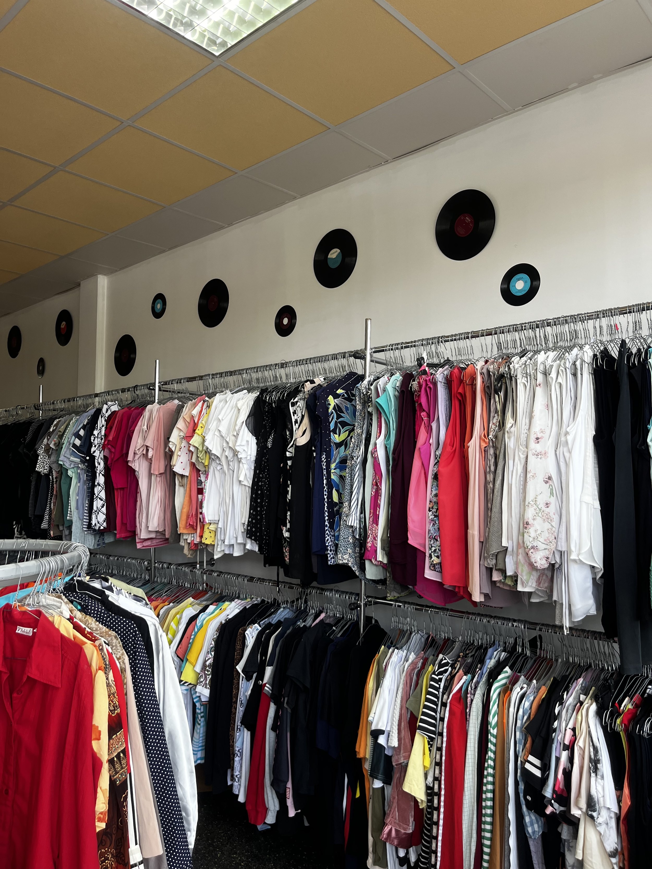 Sara Second Hand in Eisenhüttenstadt, Second Hand & Vintage Mode für alle. Kleidung, Haushaltsartikel & Accessories. Ladengeschäft mit Kleidung auf Kleiderstangen, bunte Hemden und Kleider, an einer Wand hängen Vinyl-Schallplatten,