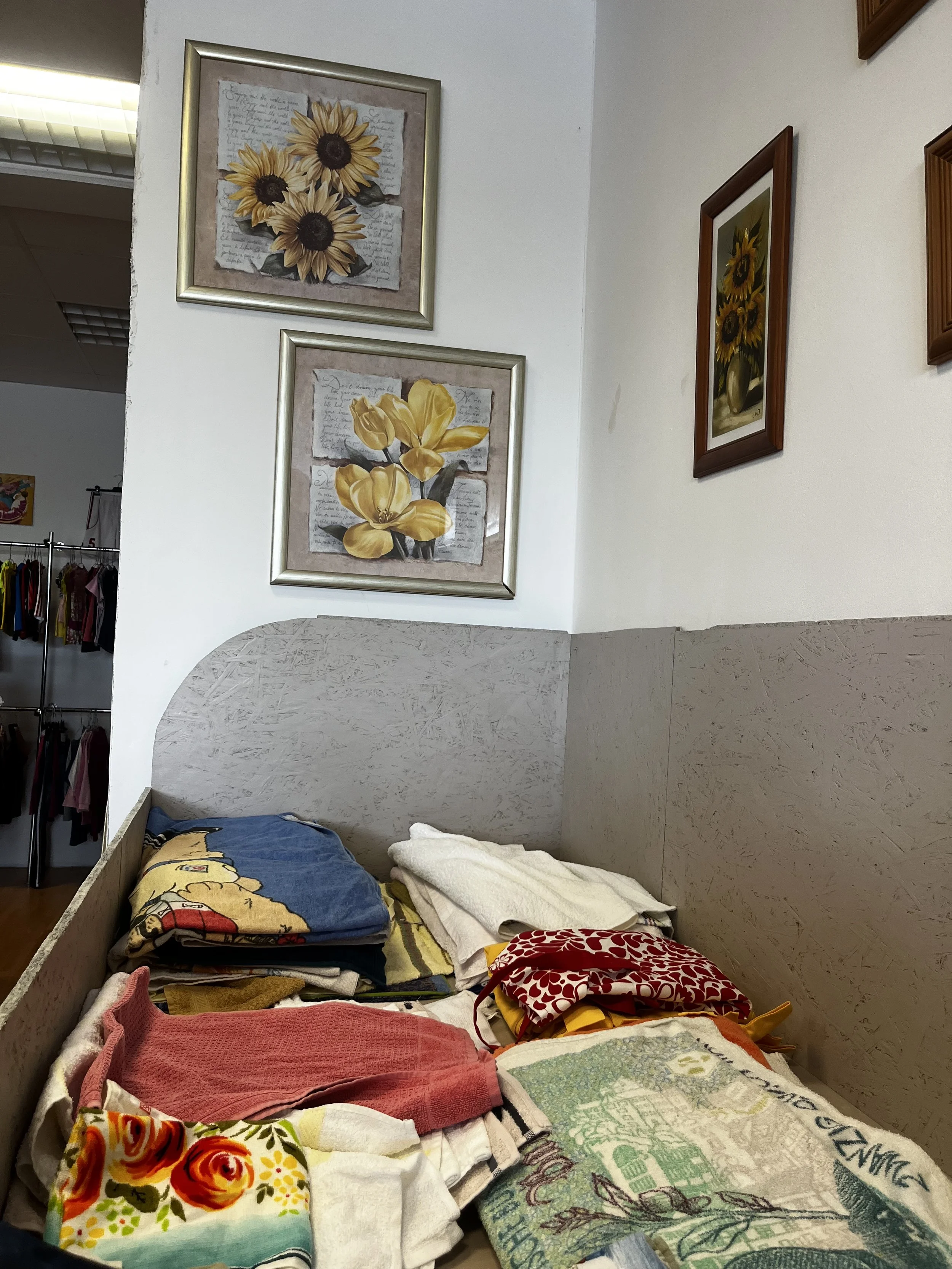 Sara Second Hand in Eisenhüttenstadt, Second Hand & Vintage Mode für alle. Kleidung, Haushaltsartikel & Accessories. Grabbelkiste mit zusammengelegten Handtüchern, Decken und Vorhängen. An der Wand hängen vier Bilder mit Sonnenblumenmotiven