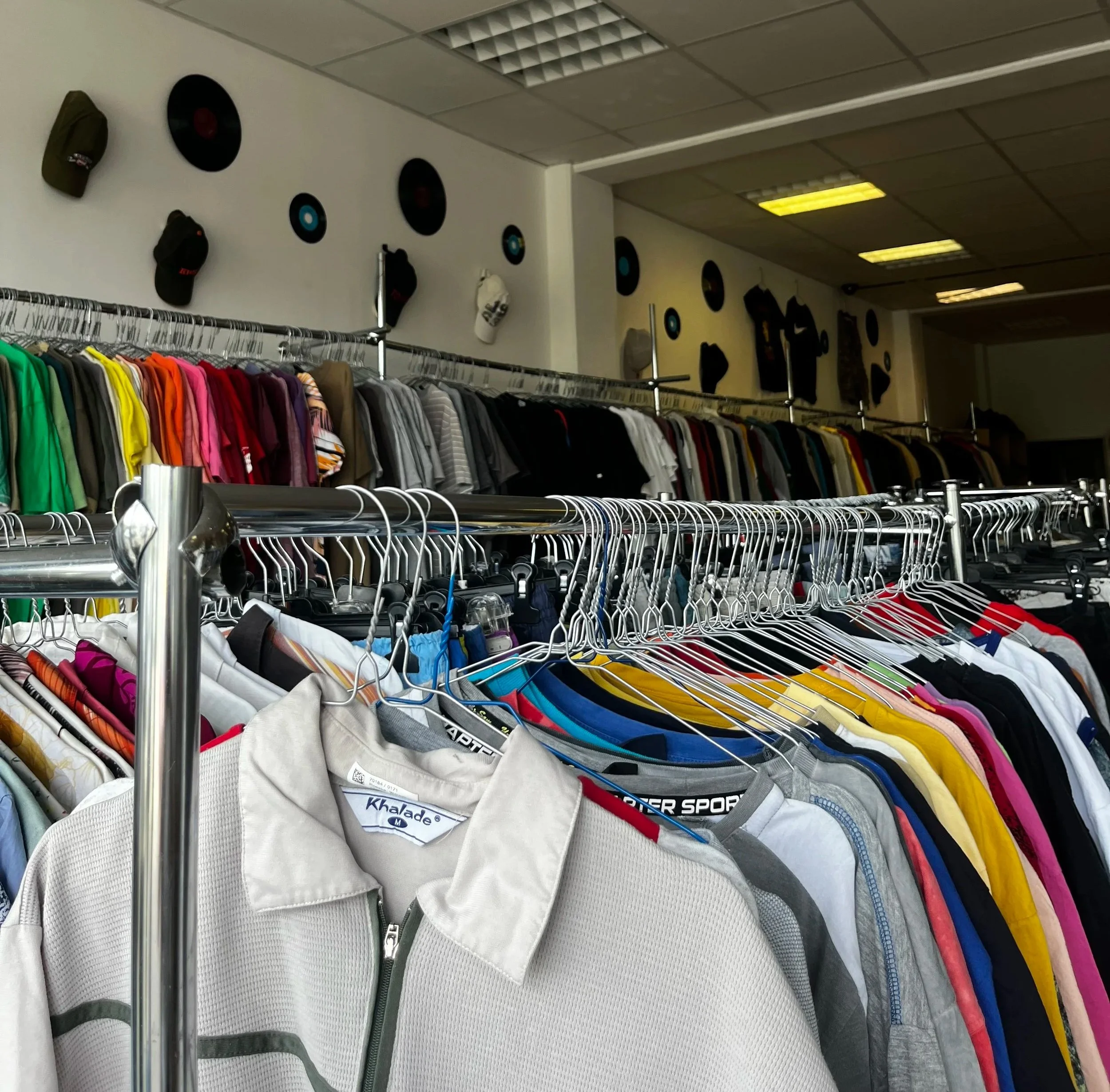 Sara Second Hand in Eisenhüttenstadt, Second Hand & Vintage Mode für alle. Kleidung, Haushaltsartikel & Accessories. Kleider und T-Shirts auf Kleiderstangen in einem Second-Hand-Shop, mit Wanddekoration aus Schallplatten und Hüten im Hintergrund.