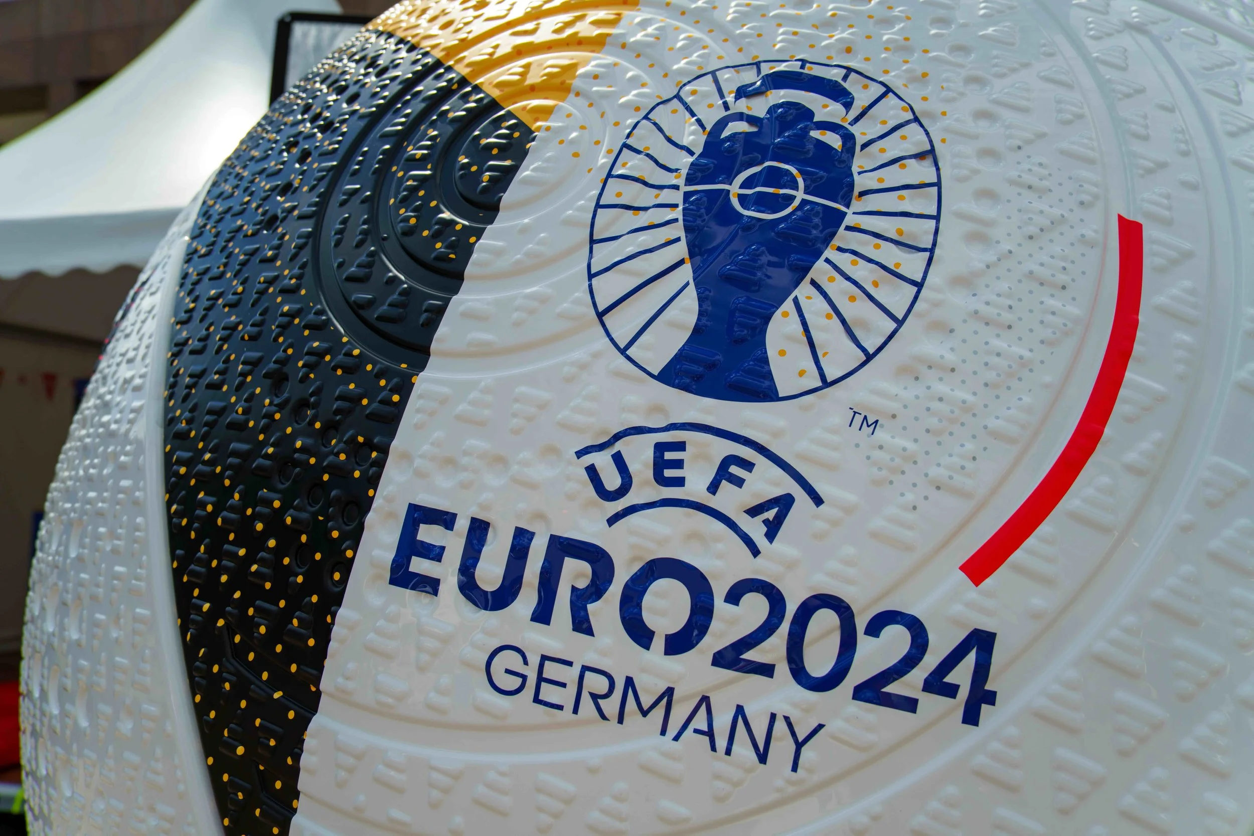 Close-up of a soccer ball with the UEFA Euro 2024 Germany logo, featuring a stylized trophy inside a circular design and the text 'UEFA EURO 2024 GERMANY' on a textured surface.