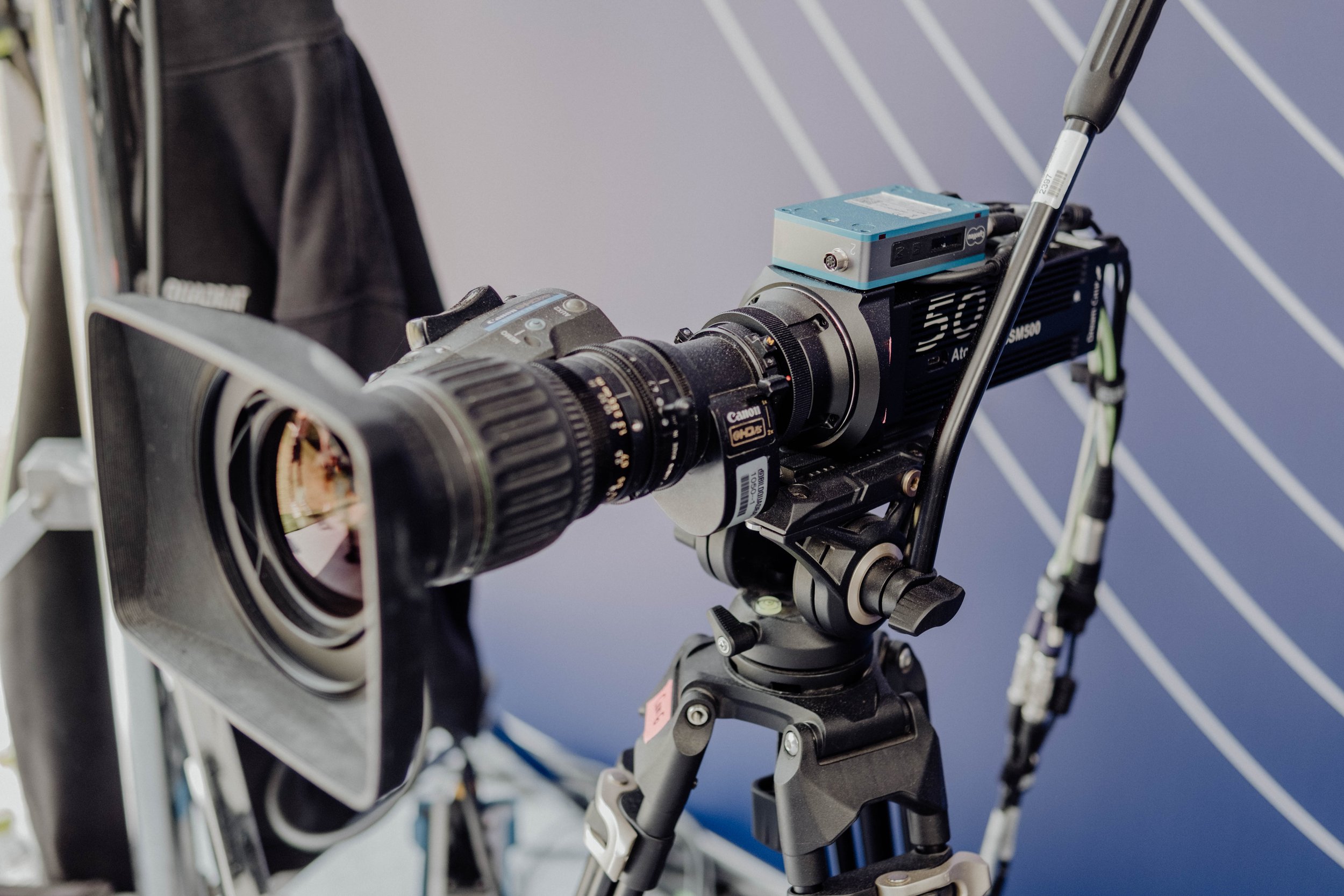A professional camera mounted on a tripod, with a large lens and a protective square filter, set up against a backdrop of blue with diagonal white lines.