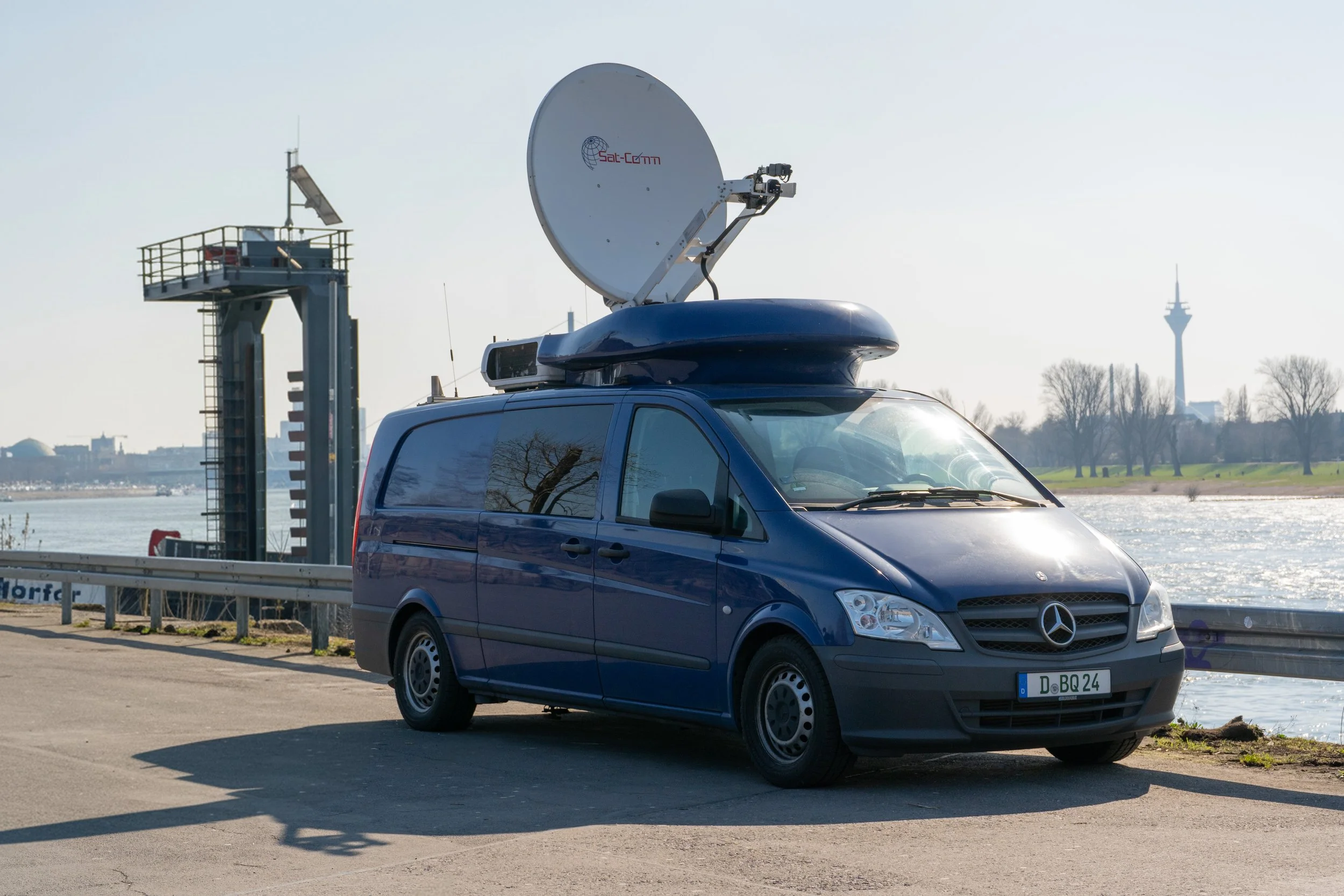 Blue Mercedes-Benz van equipped with a satellite communication dish parked near a river, with a cityscape and a TV tower in the background.