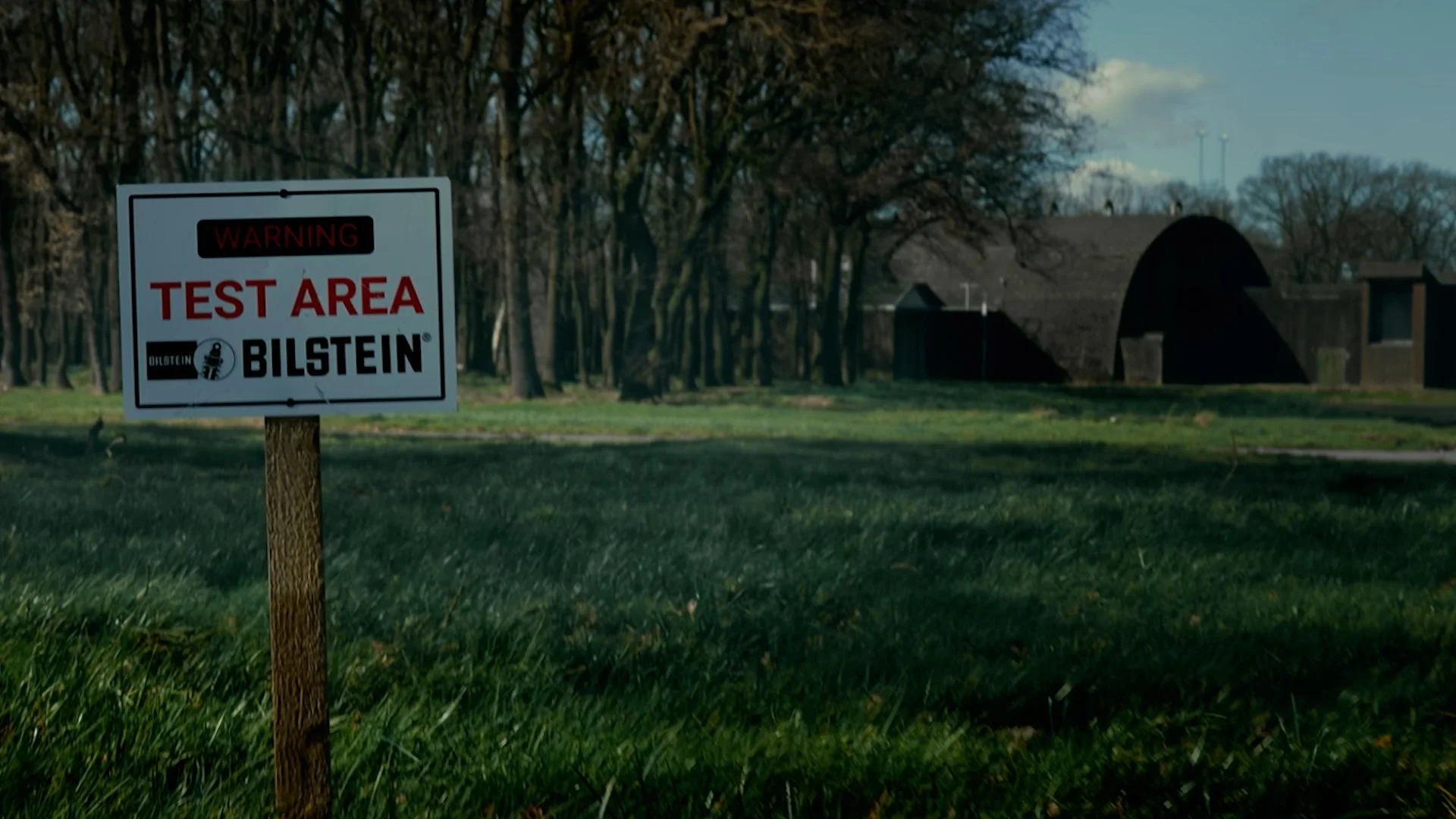 A warning sign reads 'Test Area' with the Bilstein logo, positioned in a grassy area with trees and a dark structure in the background.