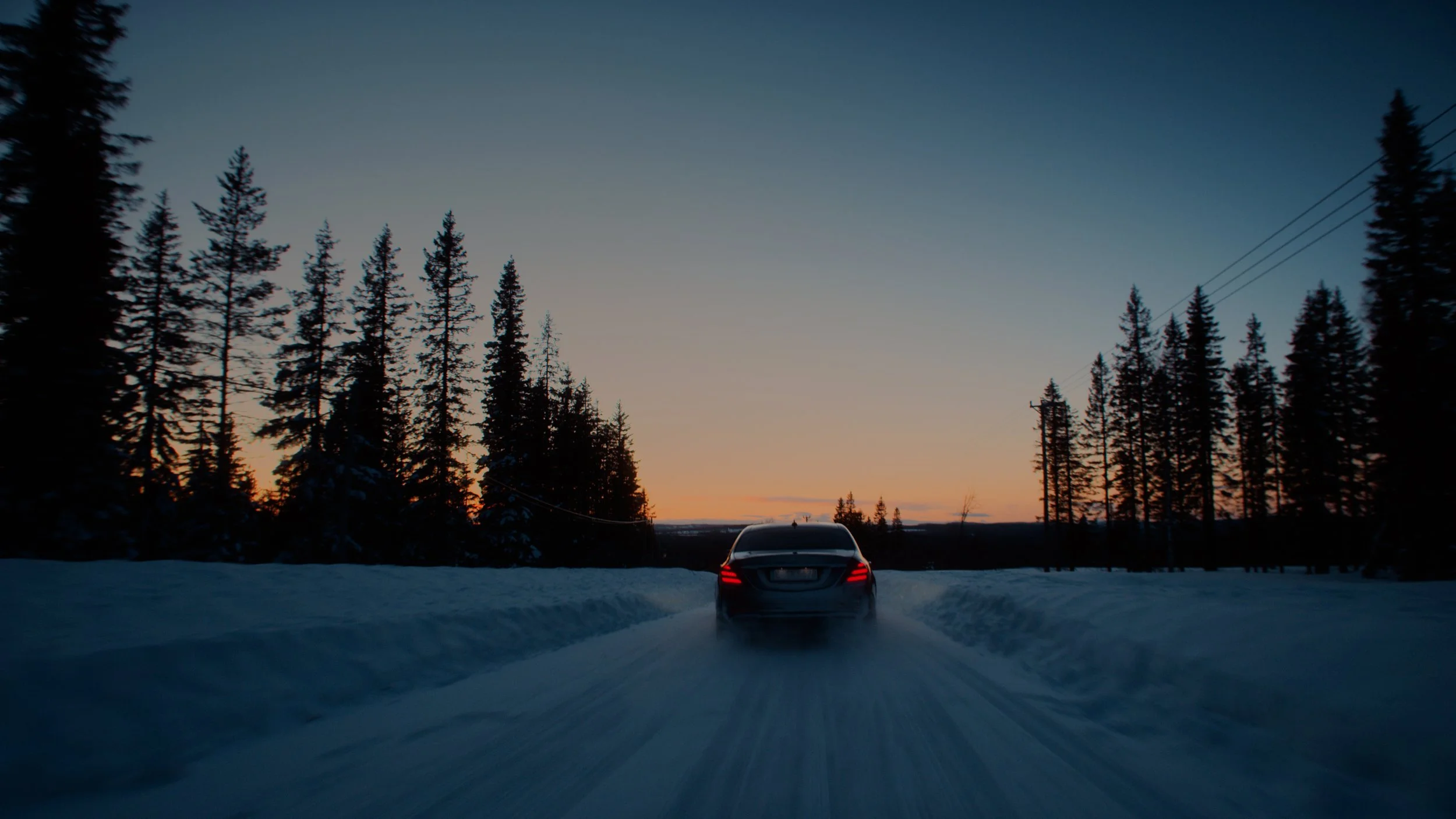 A black car driving on a snowy road surrounded by tall pine trees at sunset.