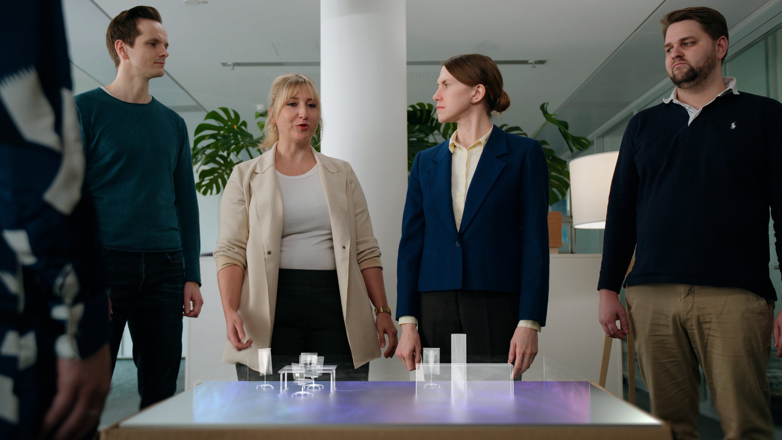 Four people standing around a table with a holographic 3D model, two women and two men, in a modern office setting with plants and lamp in the background.